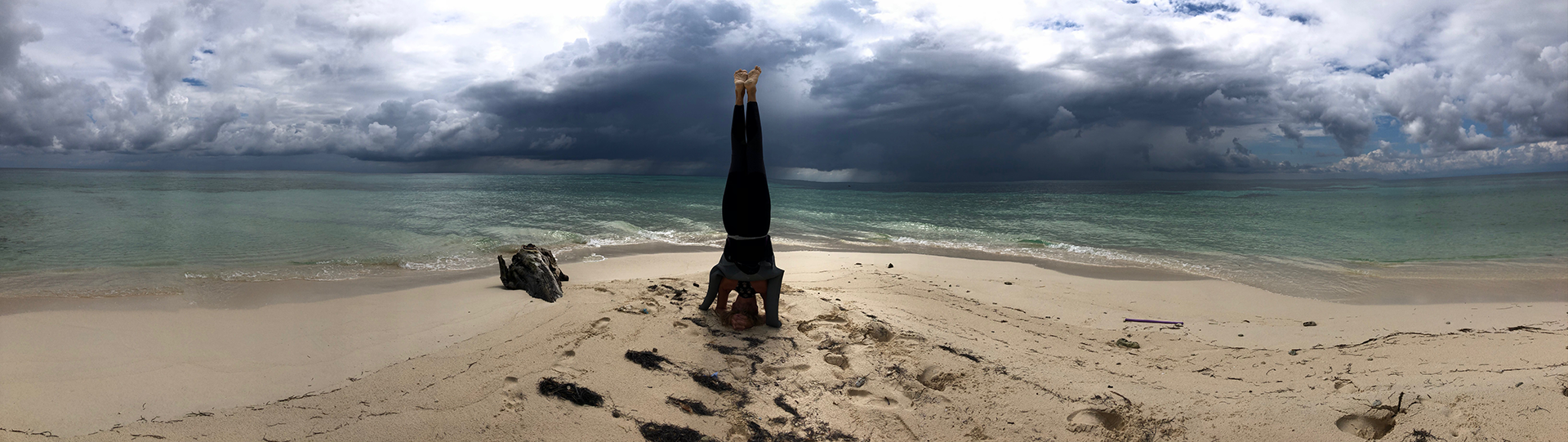 A panoramic photograph featuring a female human figure  in a headstand yoga position with her back to the camera, facing the ocean, on a beach with fine yellowish sand, calm blue green seawater, and turbulent cloudy skies