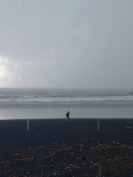 A photograph of a wide, grey, sandy beach with waves crashing in the background in overcast weather. In the middle ground, a solitary figure of a young man kicking a ball