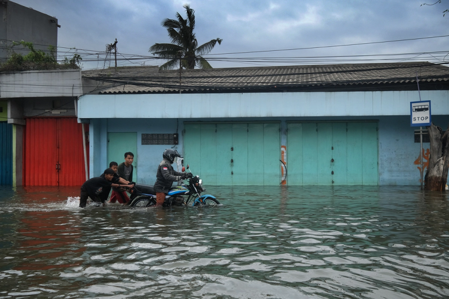 A photograph of a man wearing a bike helmet, riding atop and steering a motorbike, assisted by three young men who help push the motorbike from behind through high water that is reaching them up to their knees, suggesting a flood. In the background, brightly painted garage doors and facades of low-rise buildings, a palm tree, and a cloudy sky, suggesting a tropical setting.  