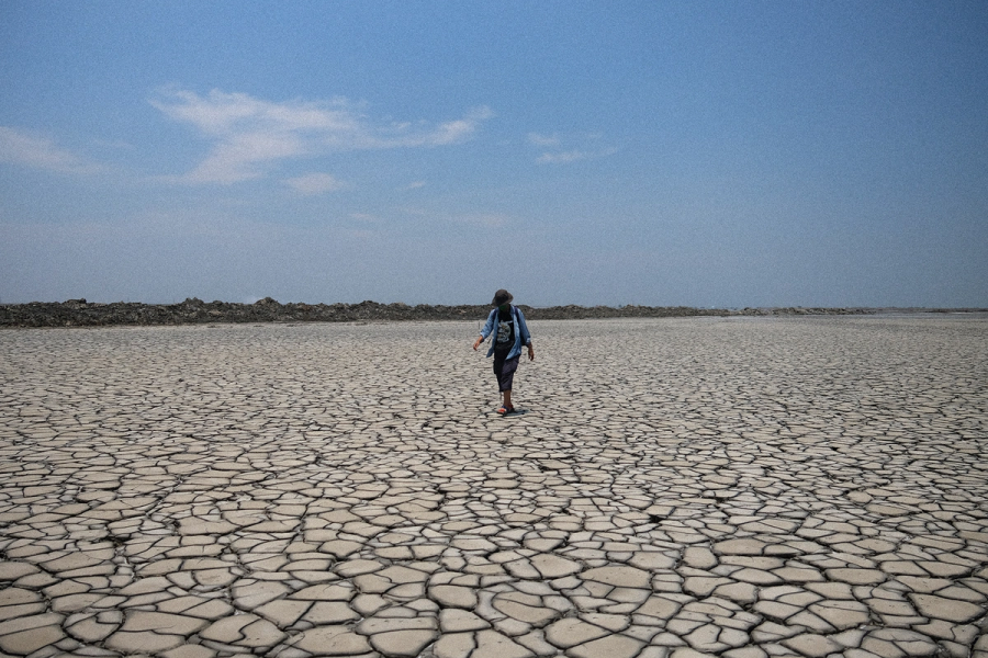 A photograph of a man wearing summer clothing and a hot, walking over a wide flat surface of cracked-up dry earth, in the far background low green vegetation and blue sky