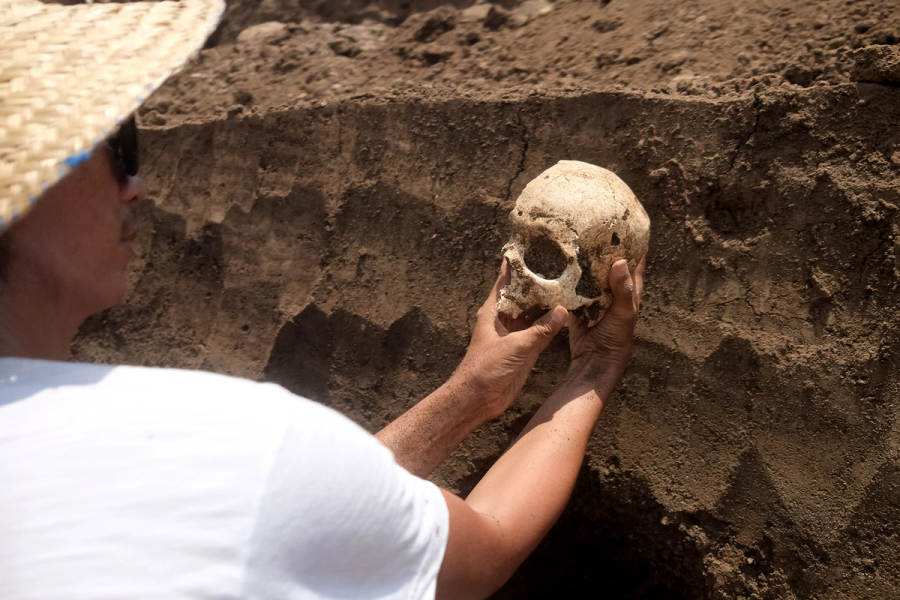 A photograph of a man wearing a straw hat and sunglasses, looking at a skull he is holding with both his hands, against the background of an earth wall.