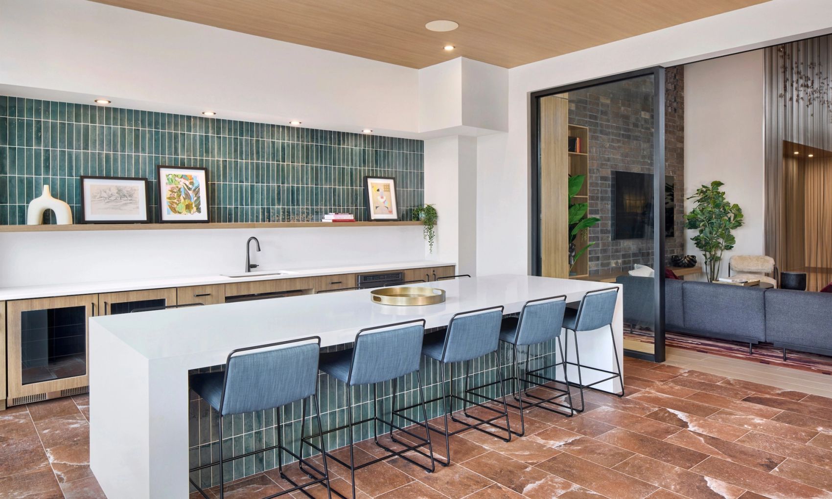 Modern resident entertainment lounge featuring a large white island with blue barstools, wood cabinetry, teal tile backsplash, and an open-concept lounge area.