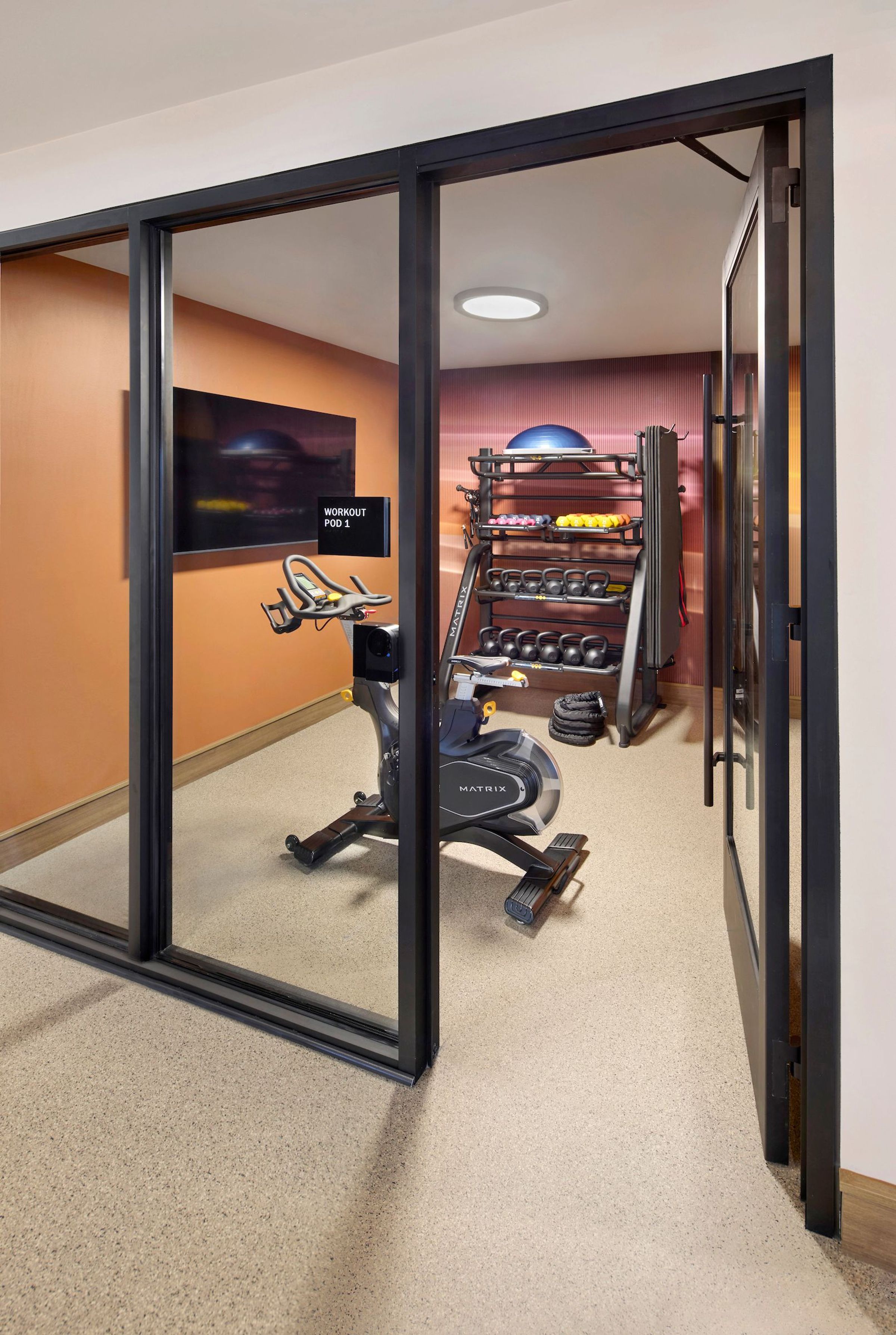 Compact fitness room with a stationary bike, free weights, kettlebells, mats, and mirrored wall.