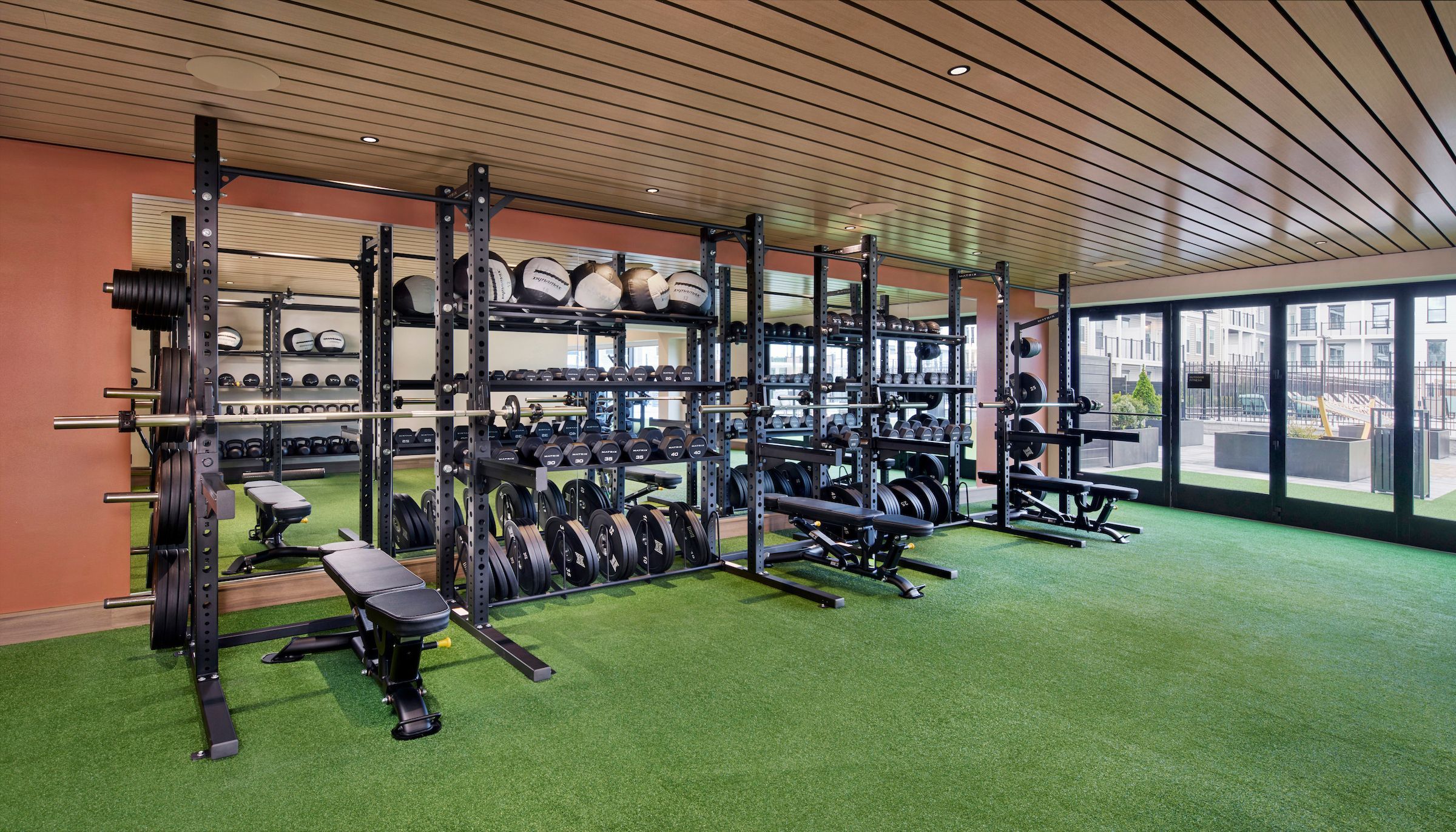Spacious fitness center featuring multiple squat racks, weight benches, free weights, medicine balls, and green turf flooring with large windows.