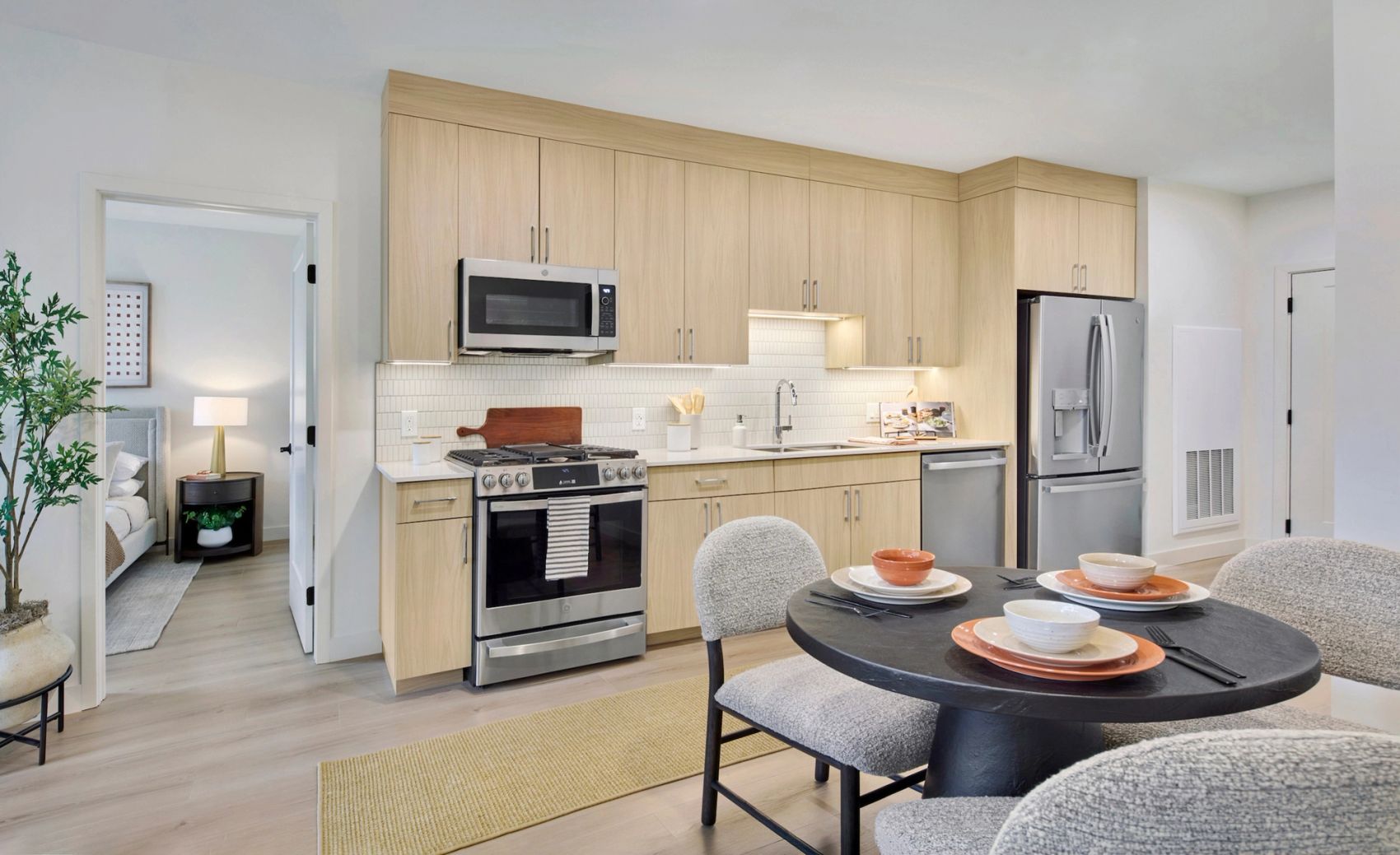 Bright apartment kitchen with light wood cabinetry, stainless steel appliances, white tile backsplash, and a round dining table set for four.