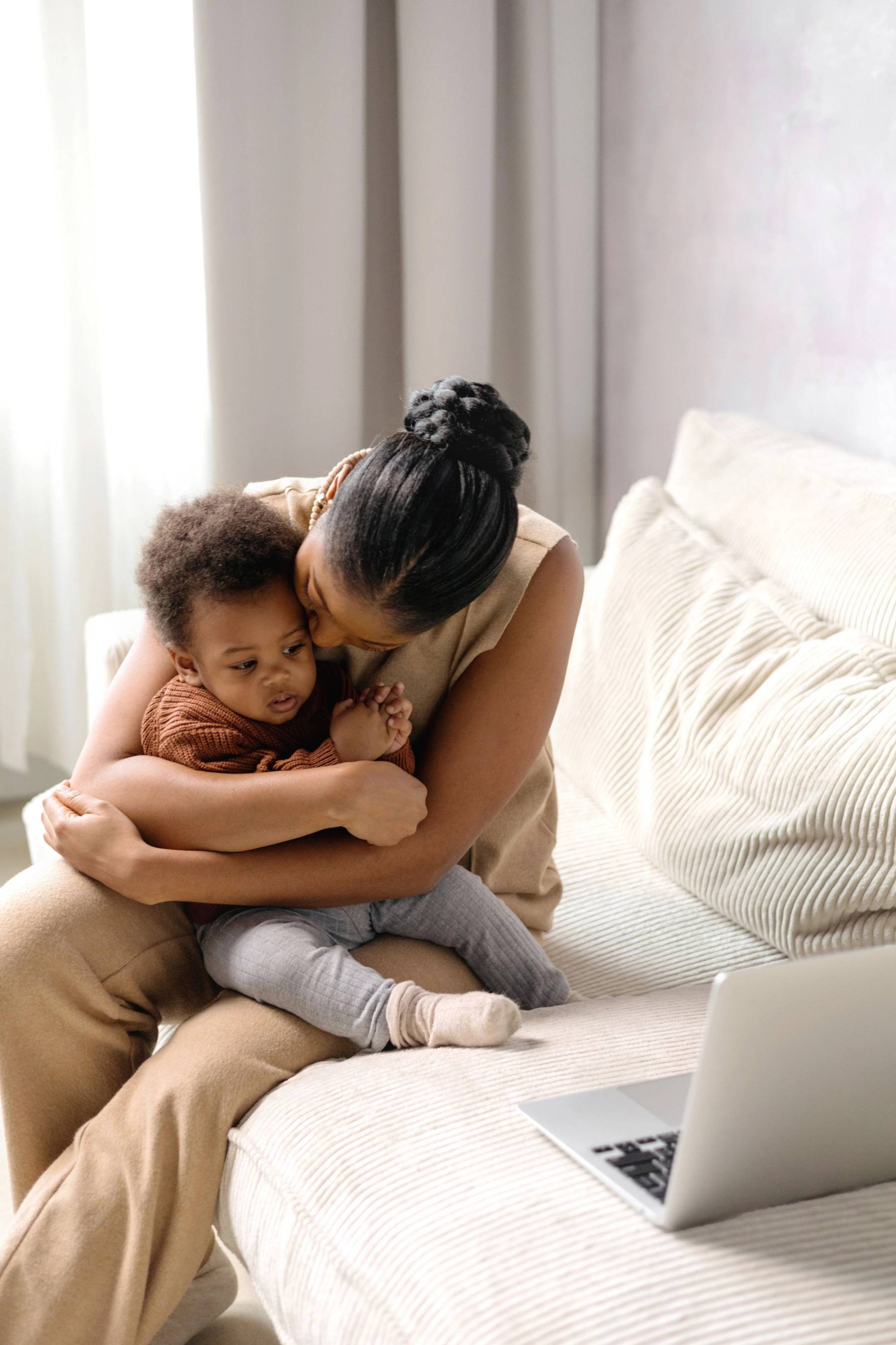 Mother lovingly hugging her child while sitting on a couch near an open laptop.