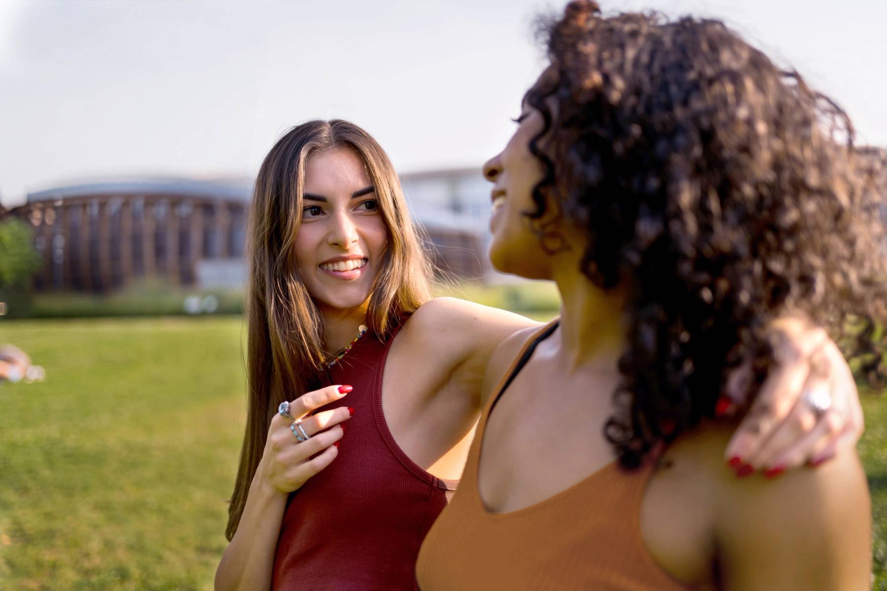 Two women smiling and talking while walking together in a sunny outdoor park setting.
