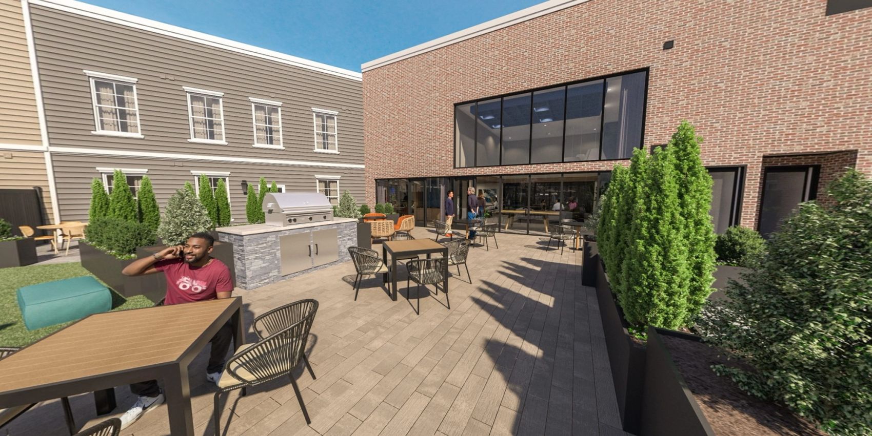Outdoor patio area with tables, grill station, greenery, and people relaxing near a brick building.