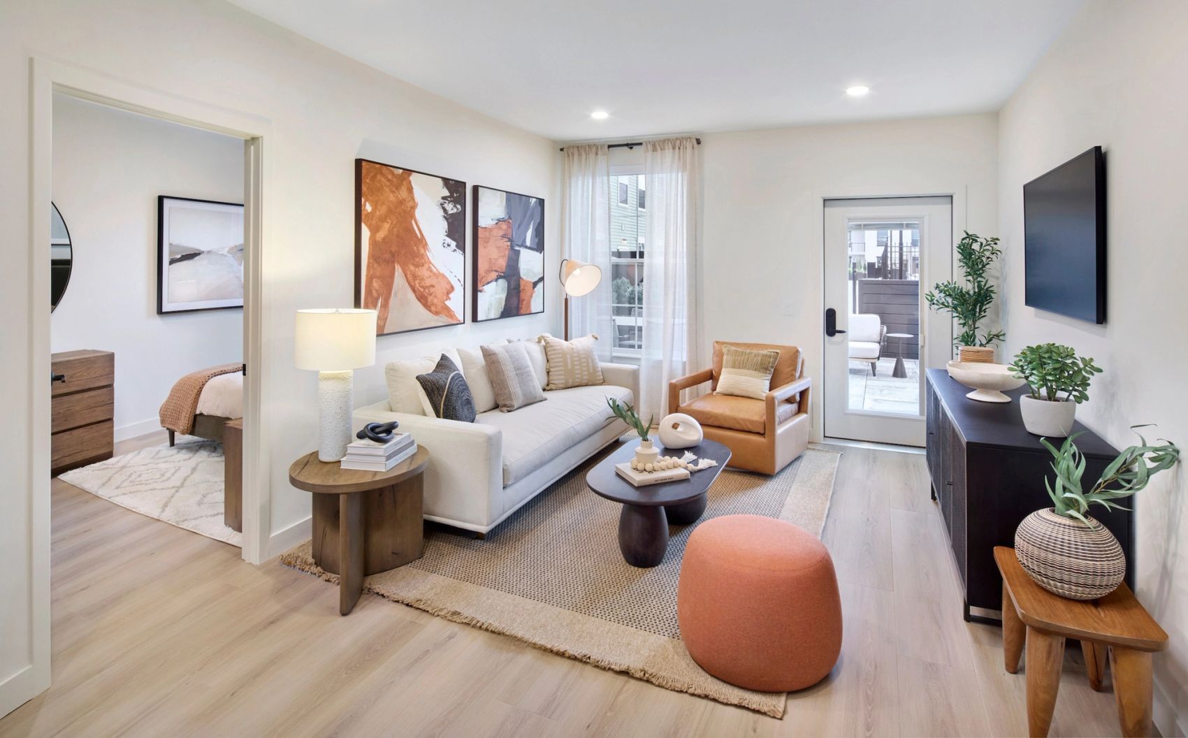 Bright apartment living room with a sofa, accent chair, coffee table, wall-mounted TV, contemporary artwork, and a glass door leading to a private outdoor space.