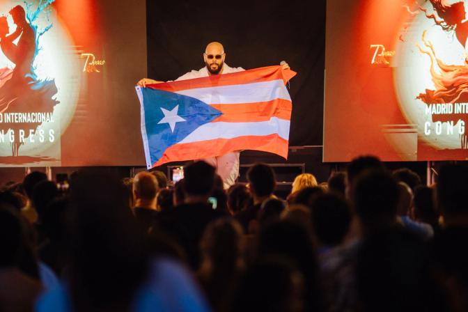 Pinto Picasso on stage holding a Puerto Rican flag before a crowd, with orange event screens and partial text in the background for Bachata. 