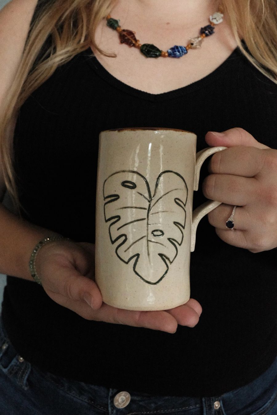 A tall cream mug hand-decorated with a monstera leaf, held in two hands