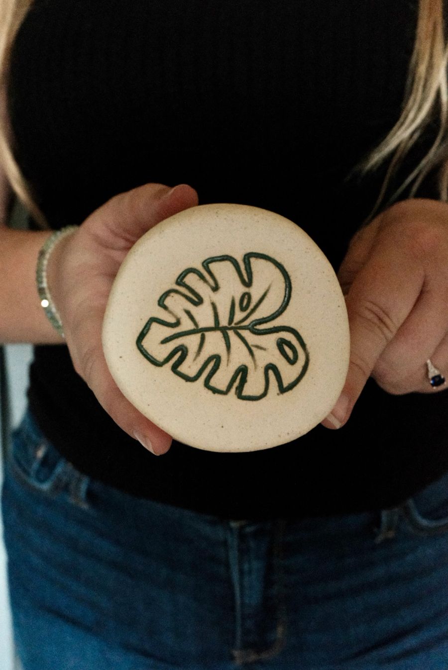Hands holding a round cream trinket dish carved with a green monstera leaf