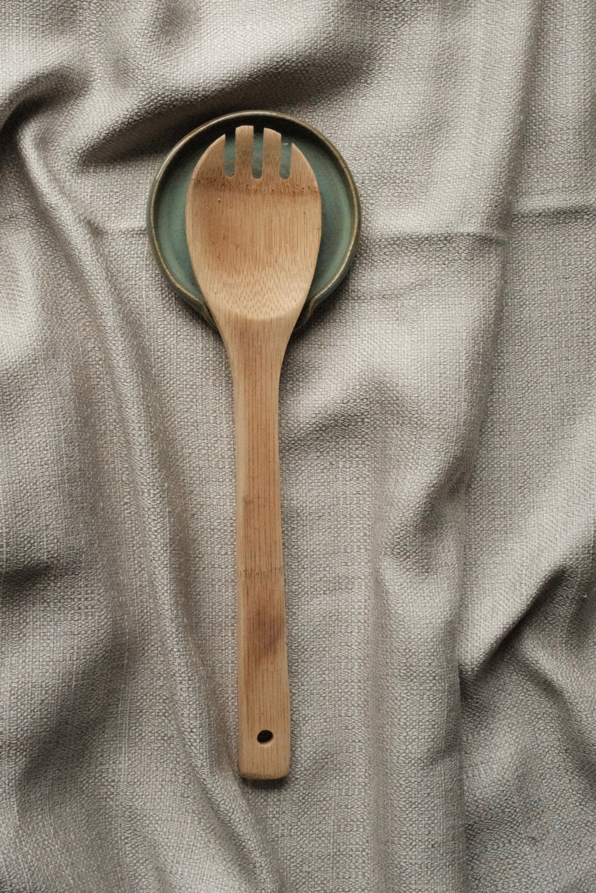 A wooden serving spoon resting on a green-rimmed ceramic spoon rest on grey linen