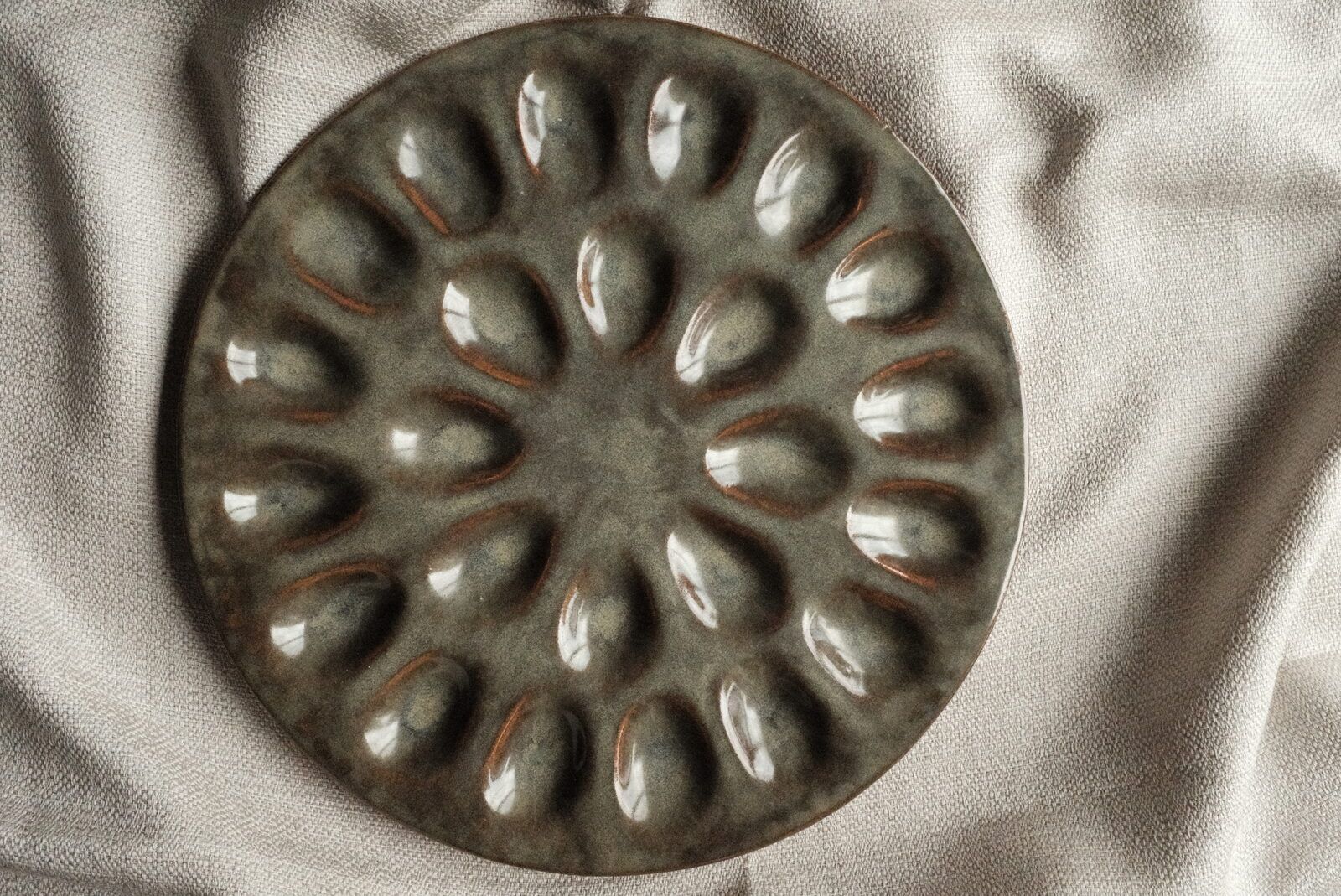 A grey-green stoneware deviled-egg plate with a petal rosette pattern, on soft linen