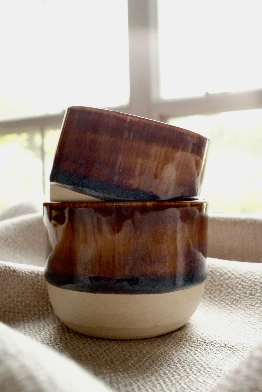 Two stacked tumblers in an amber drip glaze over raw clay, by a window