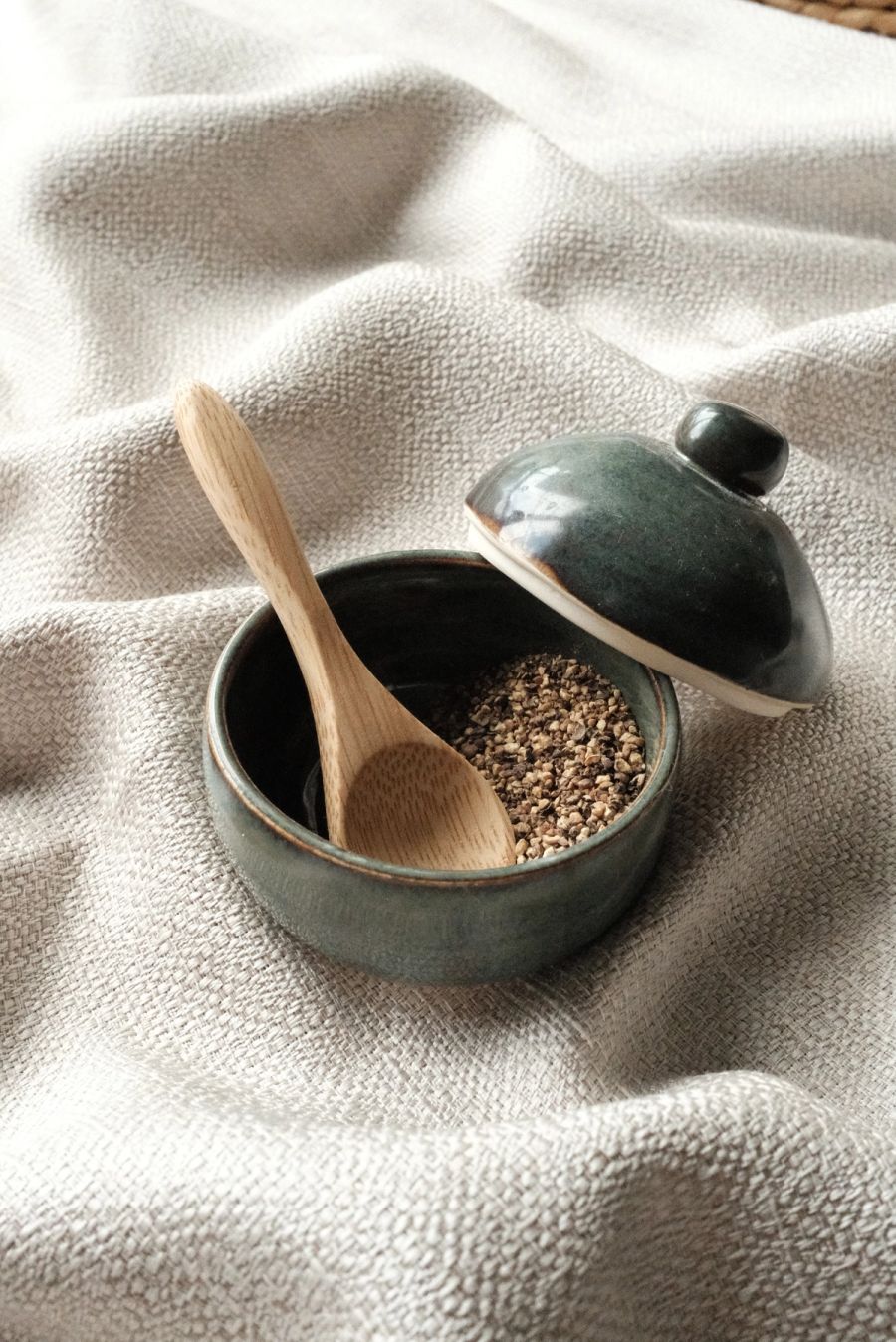 A dark teal lidded salt cellar with the lid ajar, a small wooden spoon and coarse salt inside, on linen