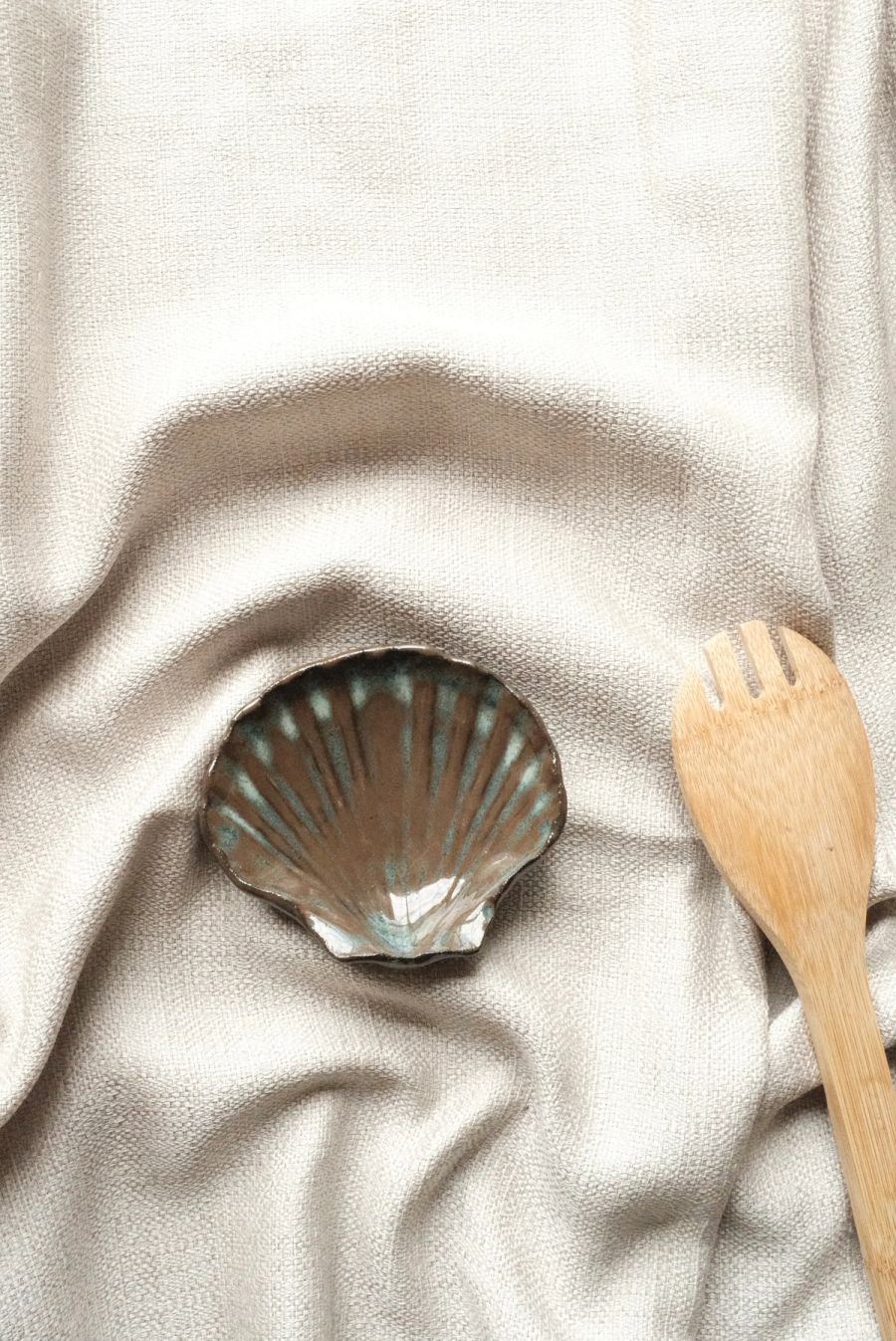 A grey-blue scallop-shell dish on cream linen beside a wooden serving fork