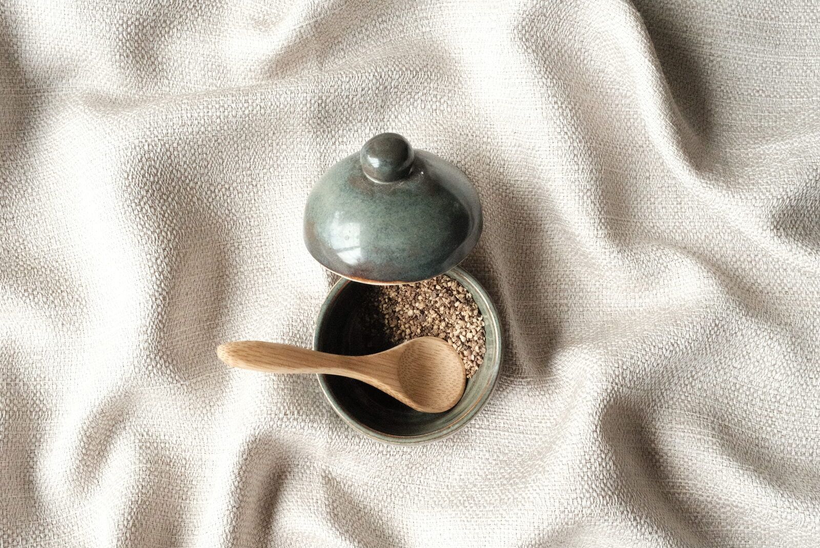A small lidded stoneware jar with a wooden spoon, resting on woven linen