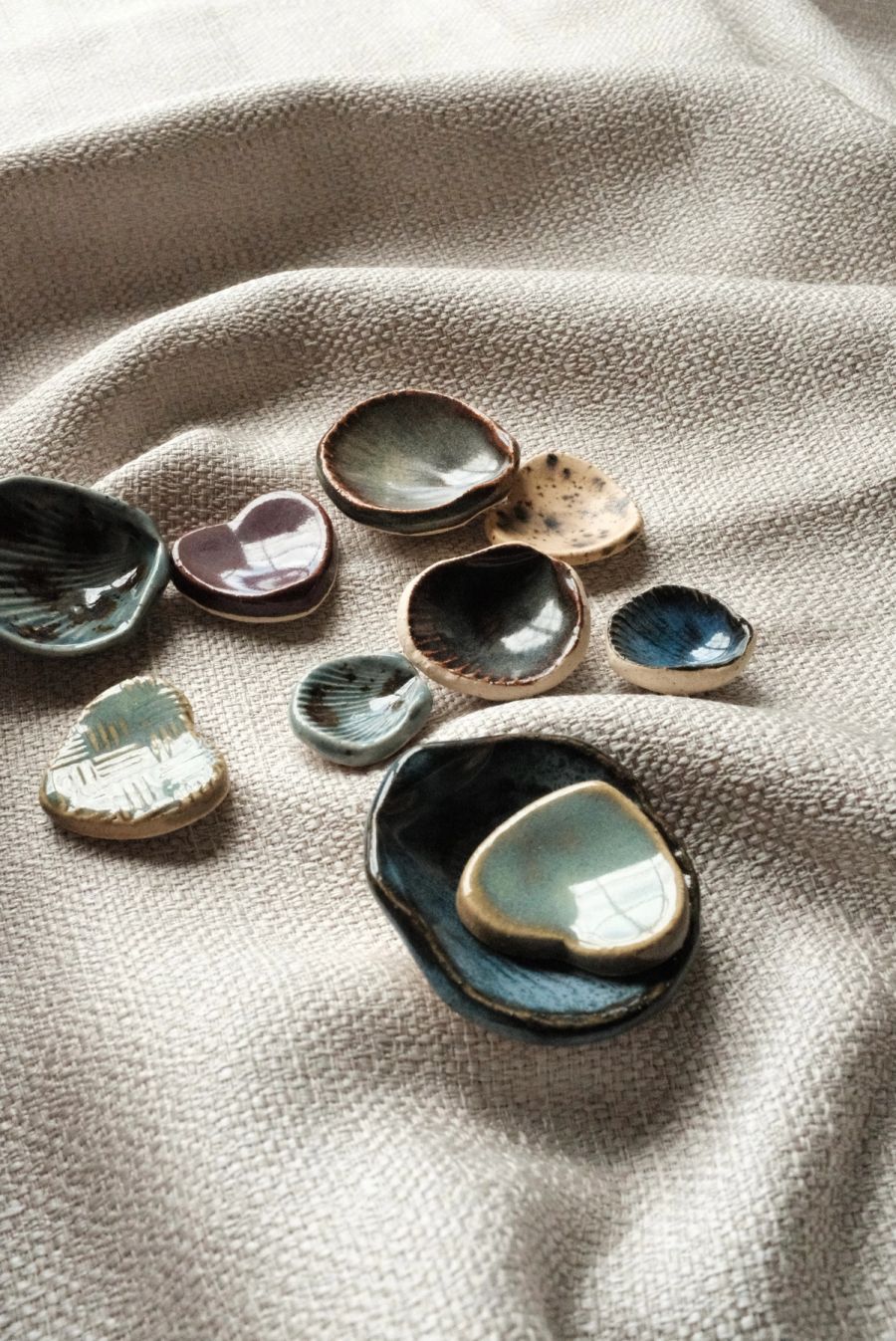 An assortment of small shell and pebble trinket dishes in blue, olive and iridescent glazes scattered on cream linen