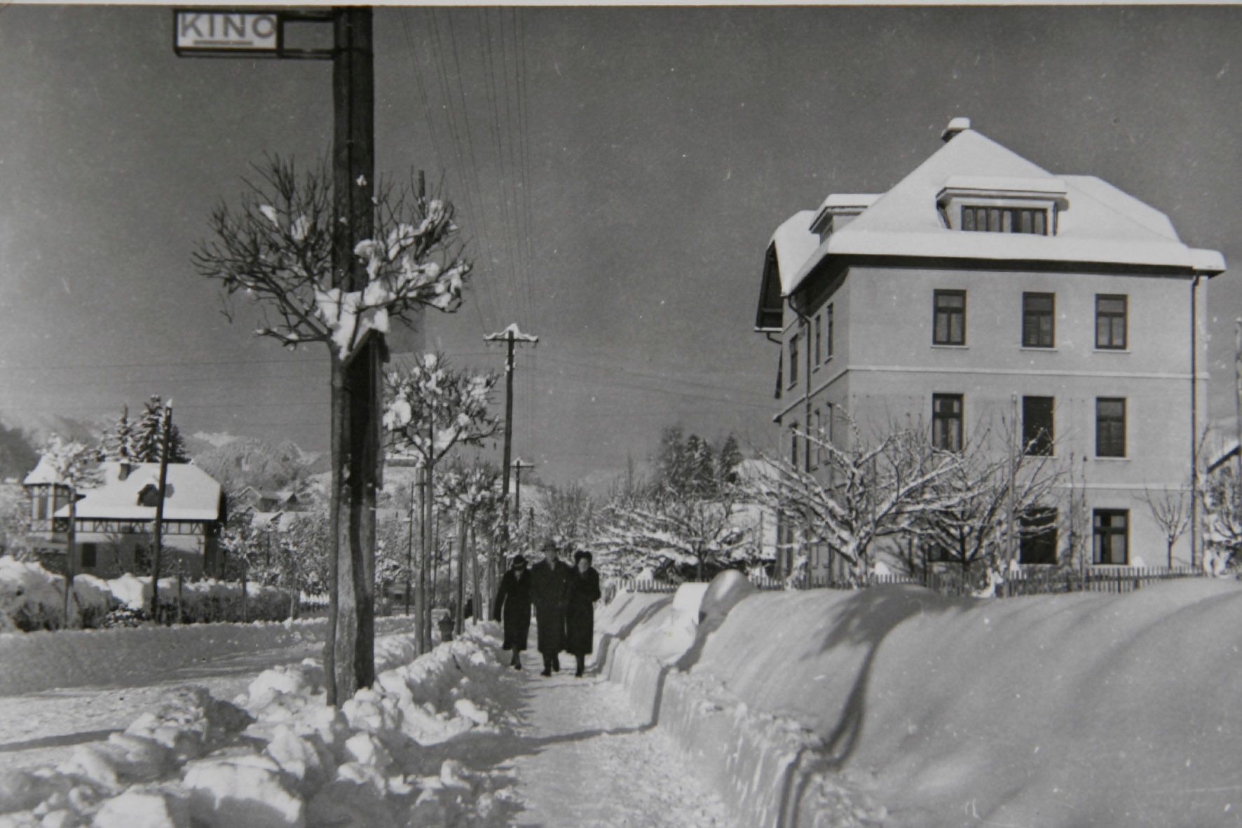 Snowy street overlooking the Josephine Villa in Bled, covered in snow, with three people walking towards the historic village. The image captures the village's wintry charm and classic architecture.