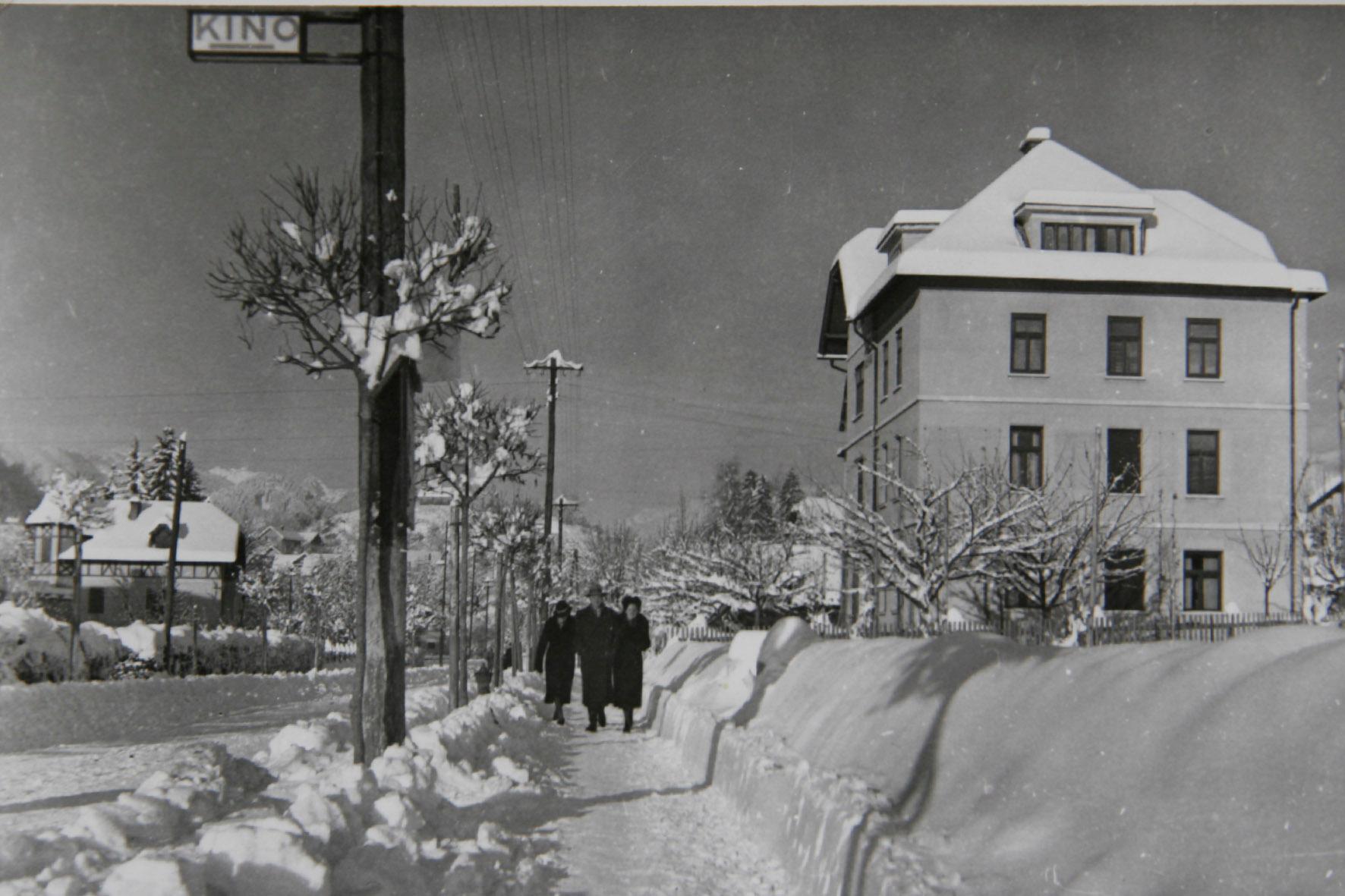Snowy street overlooking the Josephine Villa in Bled, covered in snow, with three people walking towards the historic village. The image captures the village's wintry charm and classic architecture.
