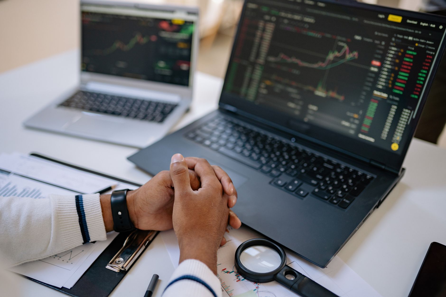 Hands clasped in front of two laptops showing market charts and a magnifier