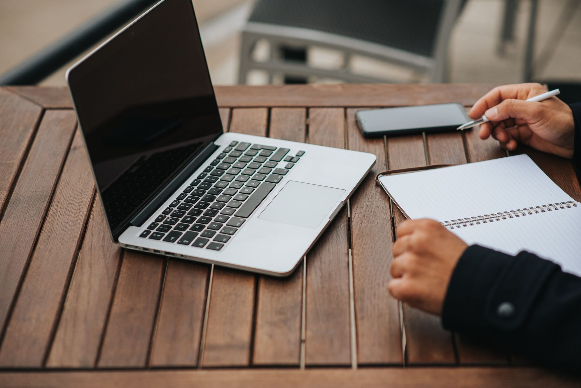 Hands writing in a notebook beside an open laptop on an outdoor wooden table