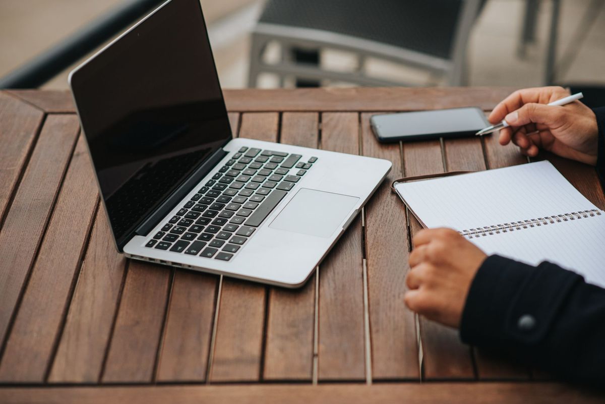 Hands writing in a notebook beside an open laptop on an outdoor wooden table