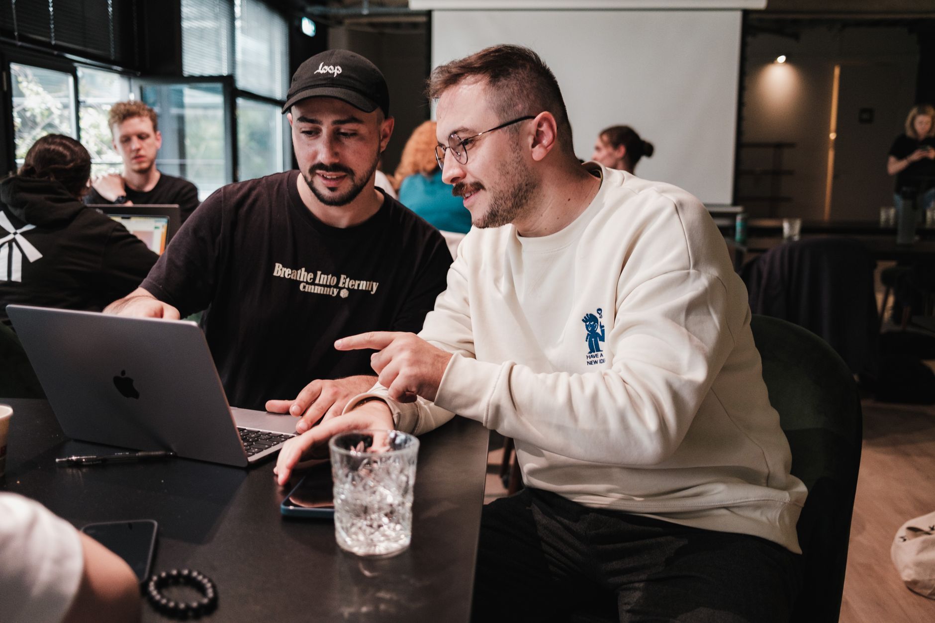 Two men reviewing something on a laptop together at a table in a busy workspace