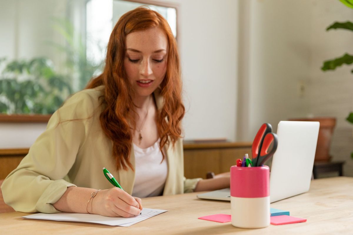 Woman writing notes by hand at a desk with an open laptop beside her