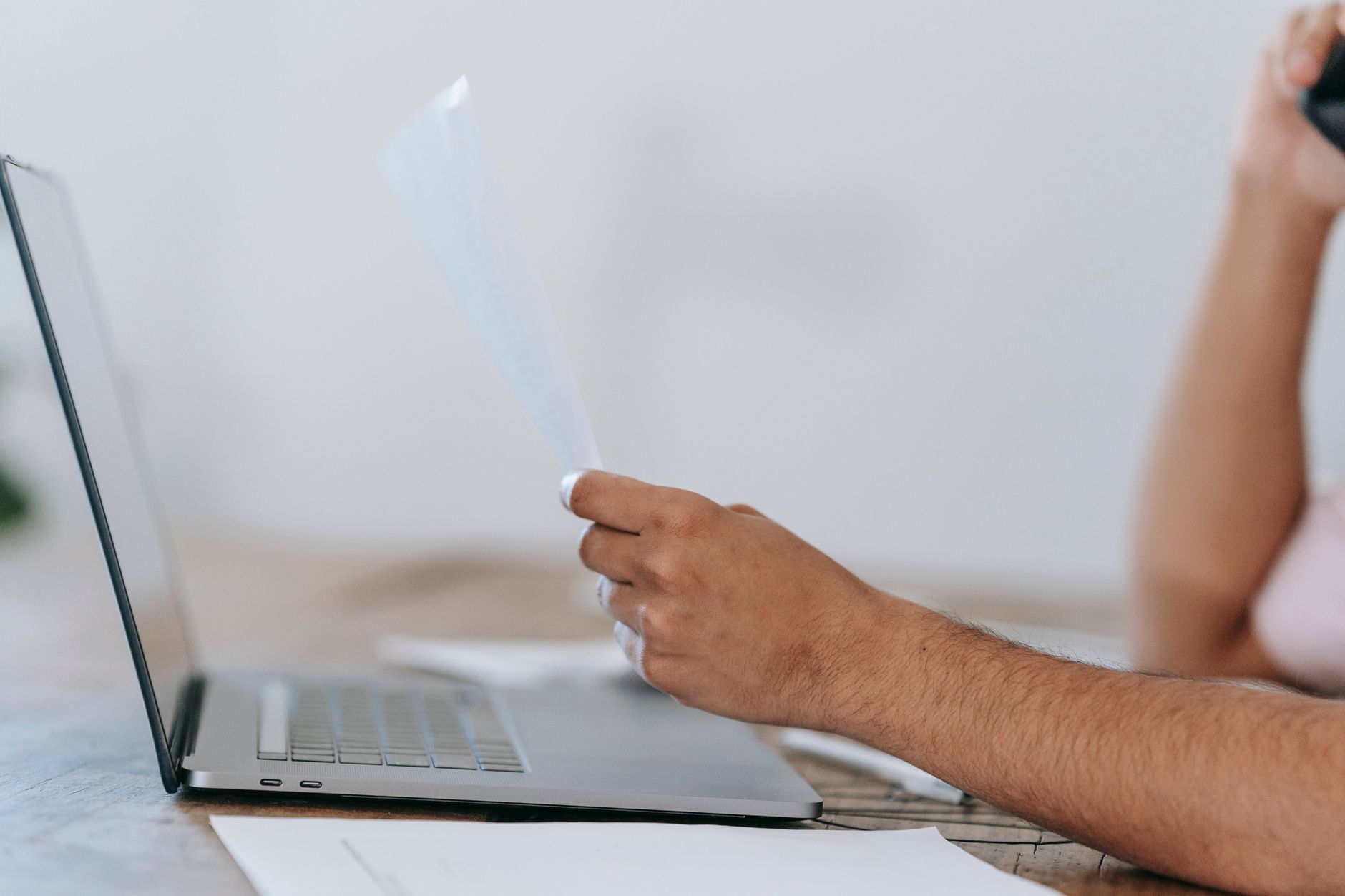 Man reading a printed page beside an open laptop at a bright desk
