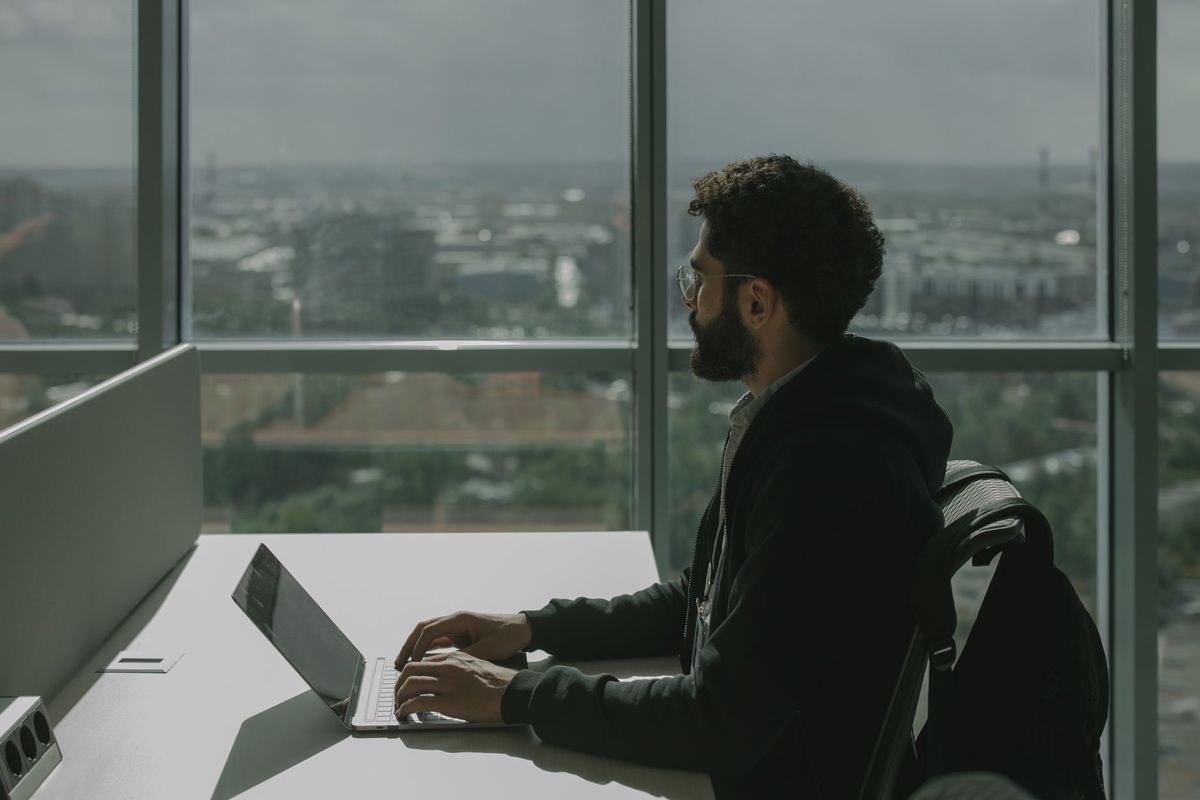 Man working alone on a laptop at a desk beside floor-to-ceiling city windows