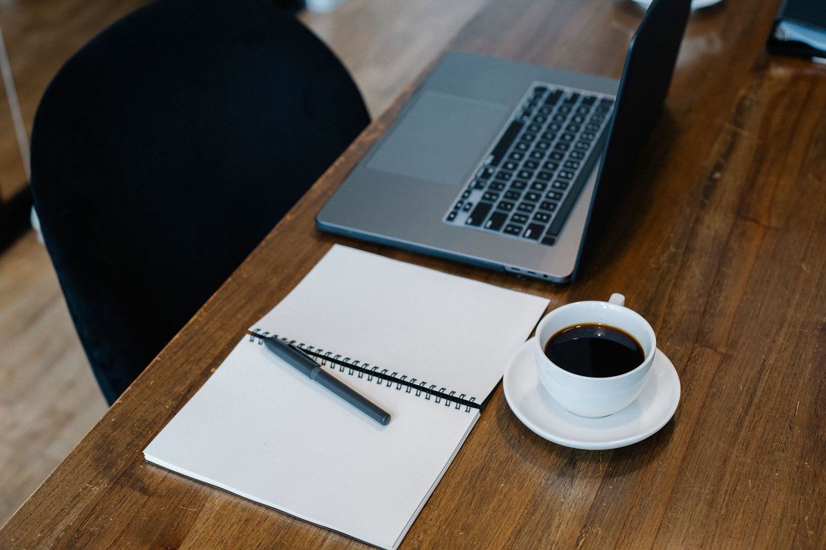 Open notebook, pen and coffee beside a laptop on a wooden desk
