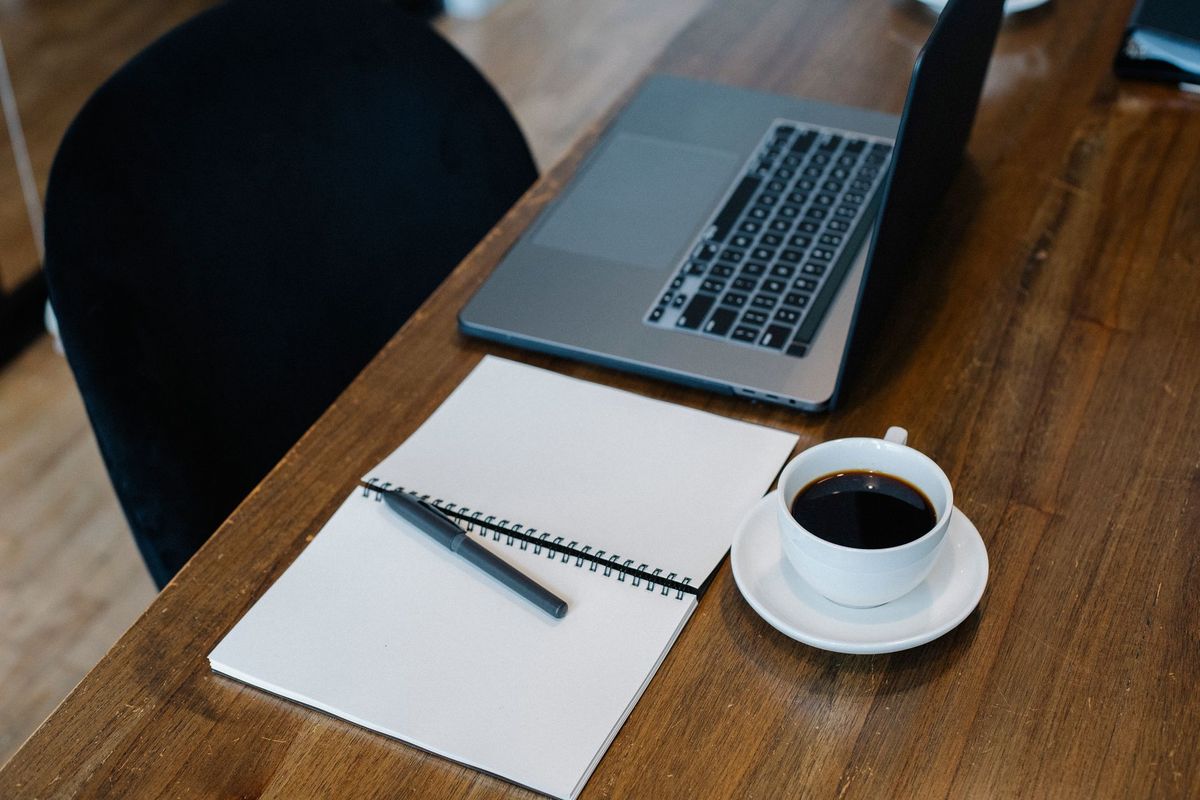 Open notebook, pen and coffee beside a laptop on a wooden desk