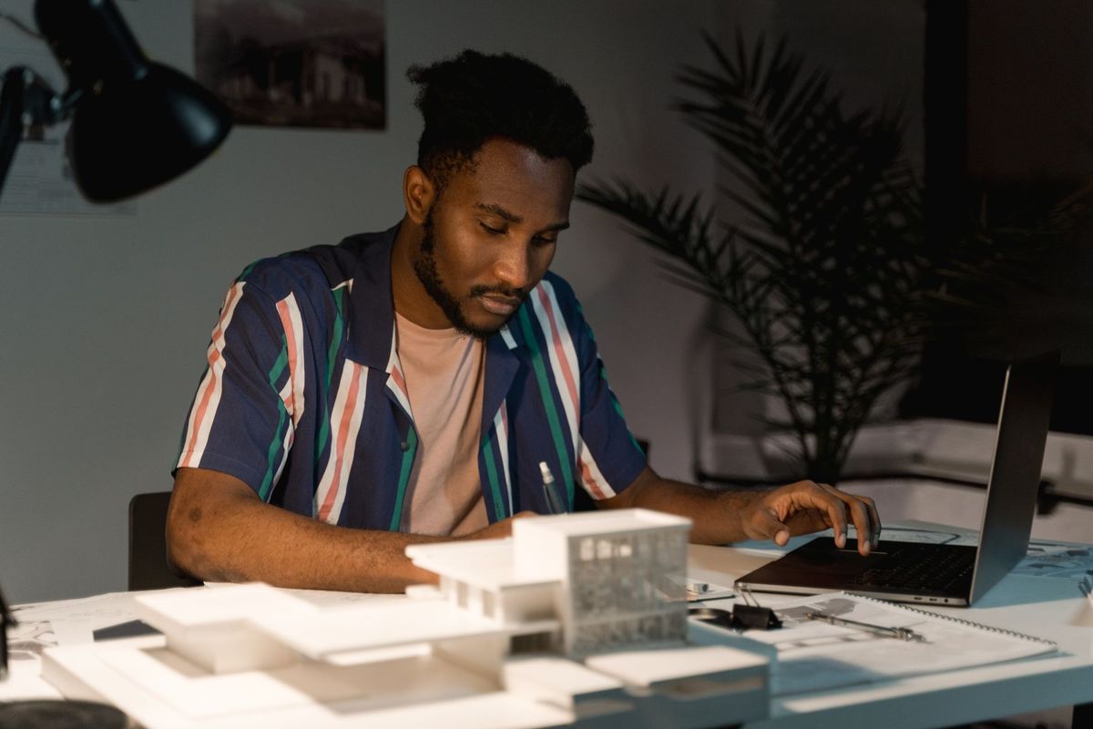 Man sketching plans at a desk with a laptop and a scale model beside him