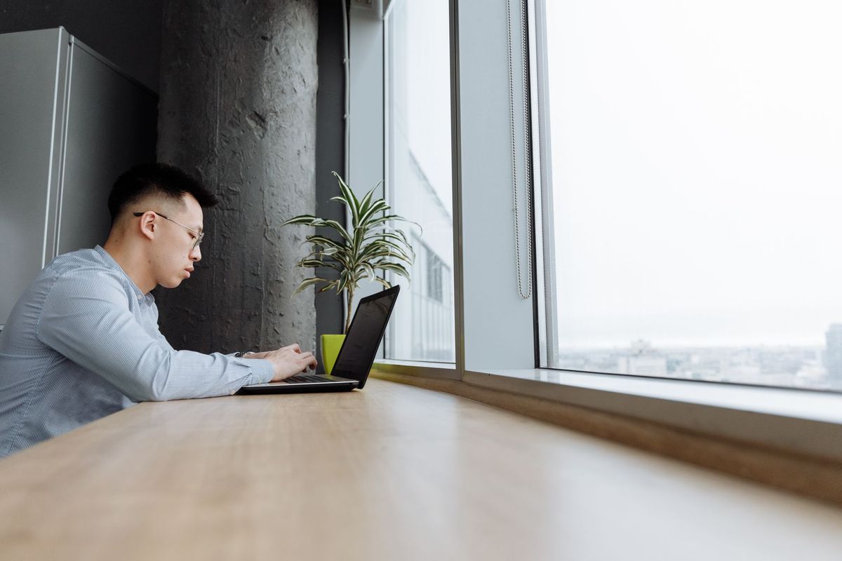 Man working on a laptop at a long wooden desk beside a bright window