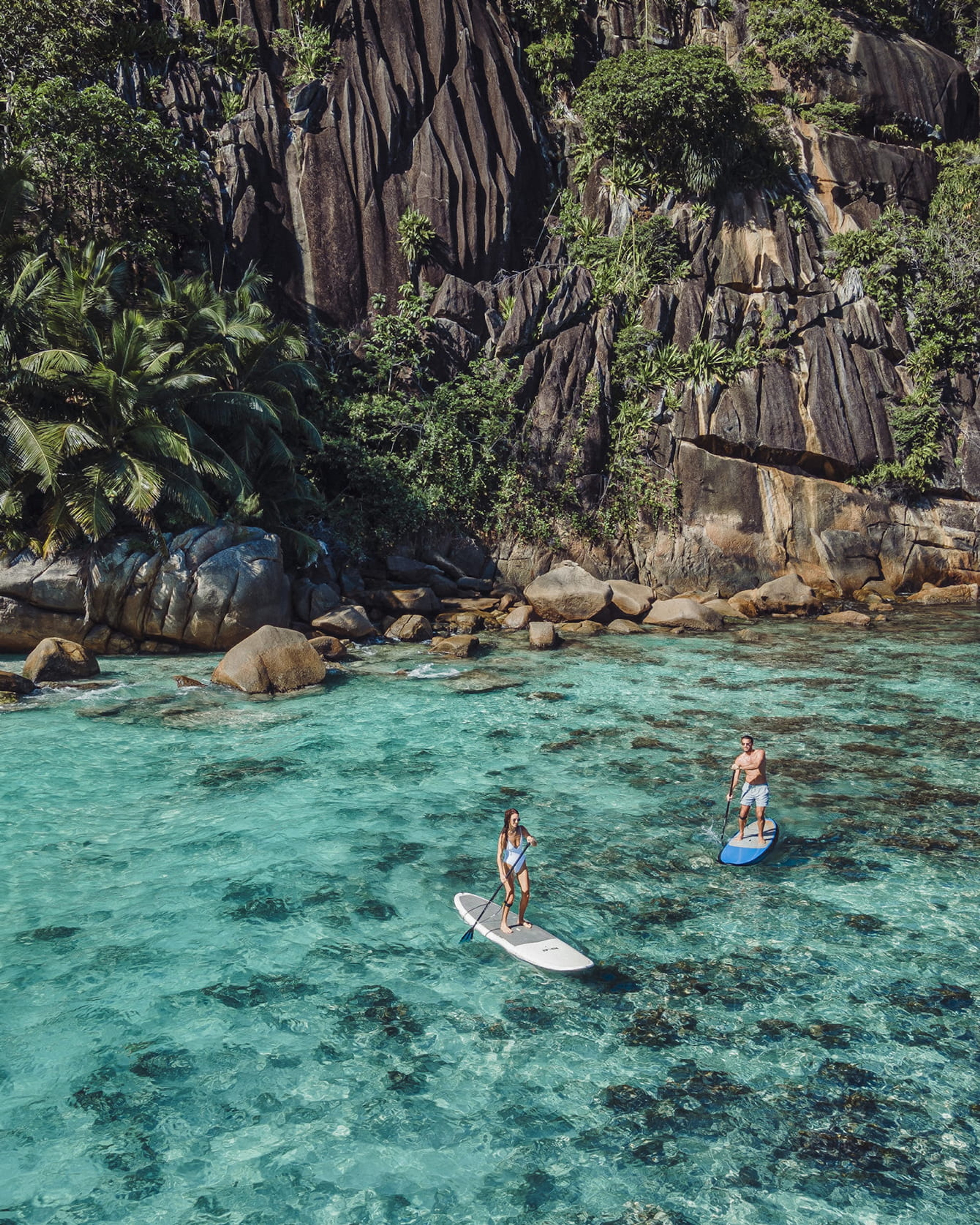 Two people paddleboarding on crystal clear water with a rocky jungle cliff in the background