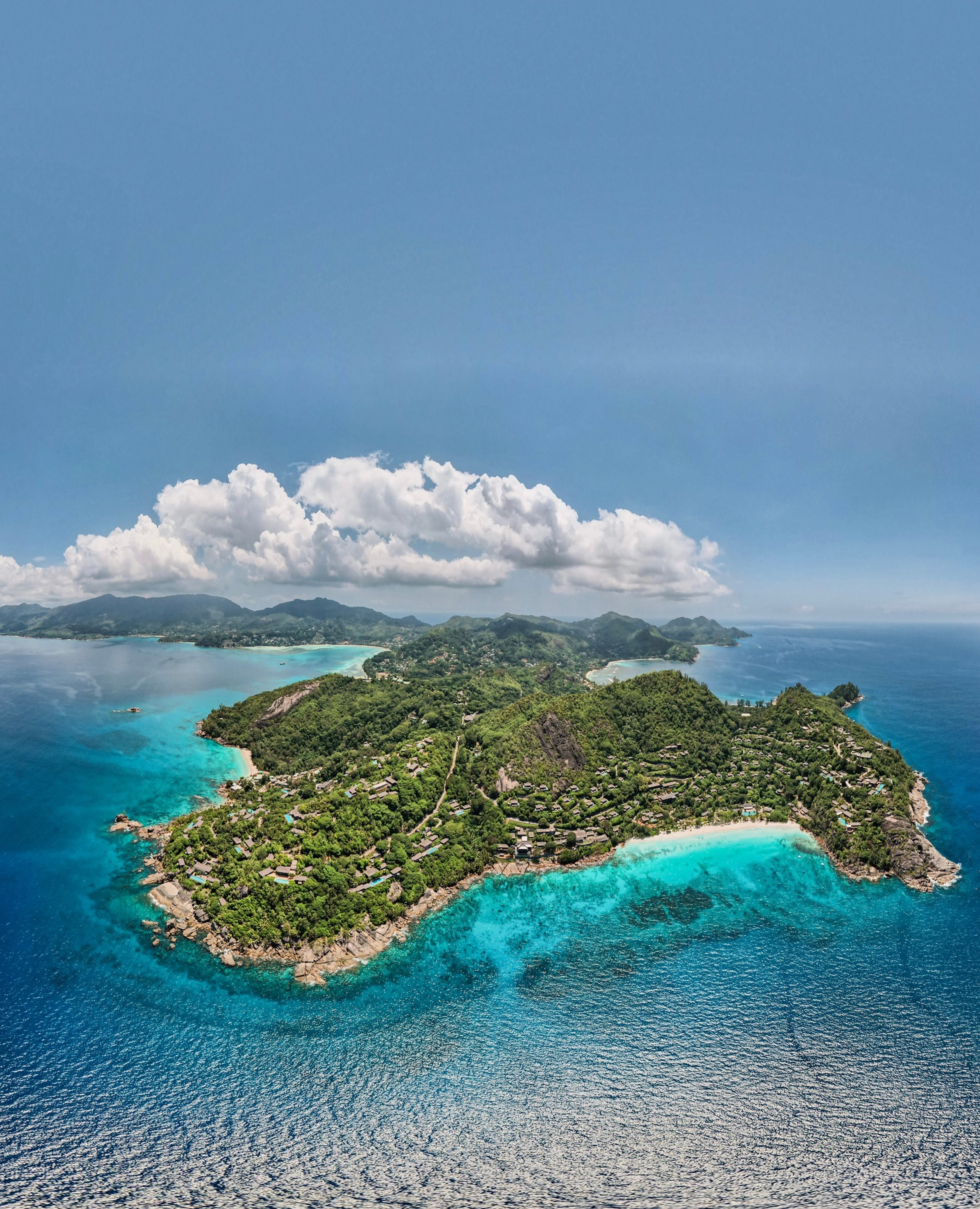 A panoramic aerial view of an island with turquoise waters, sandy beaches, and green, forested hills