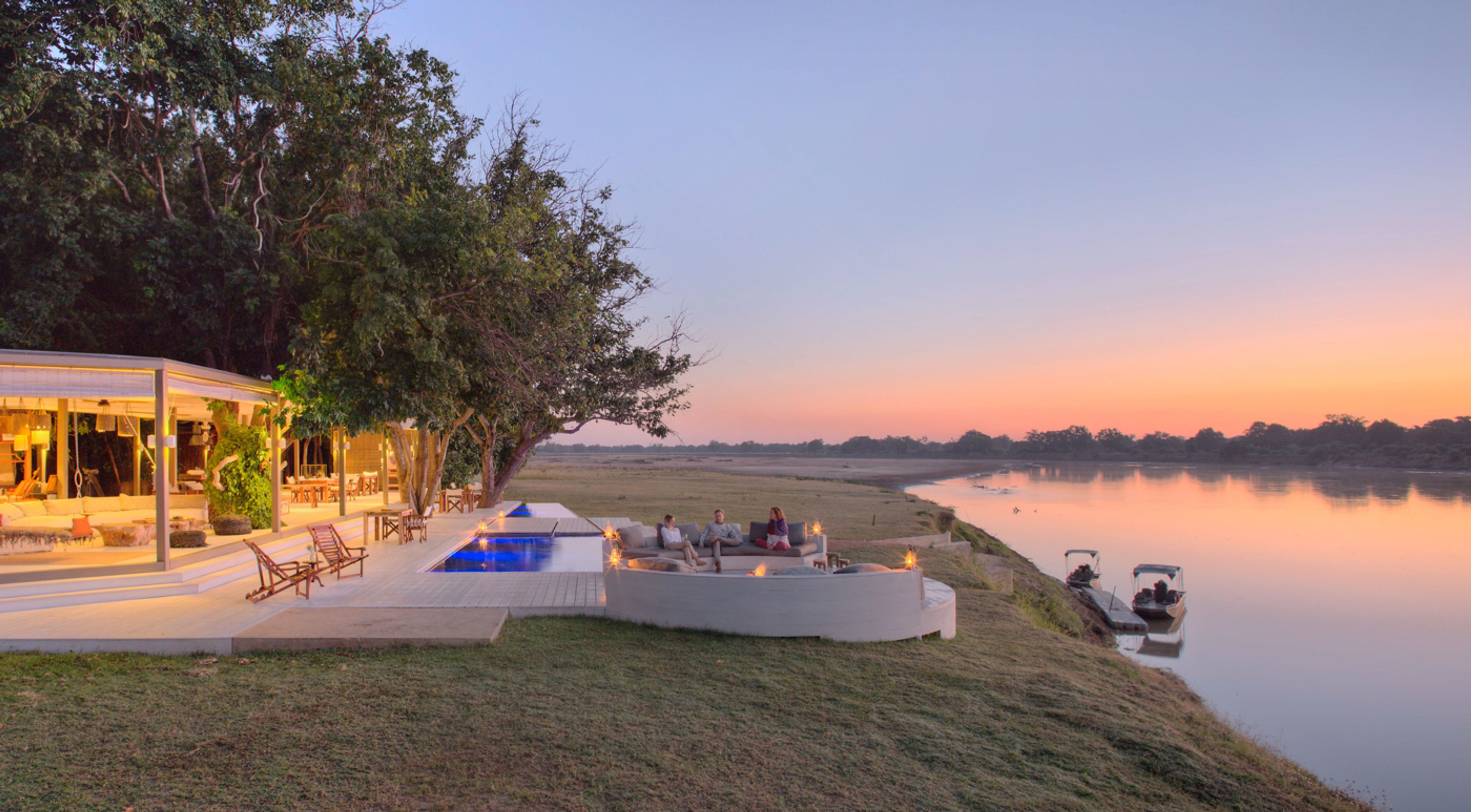 Luxury lodge by a river at sunset, with people sitting by a firepit