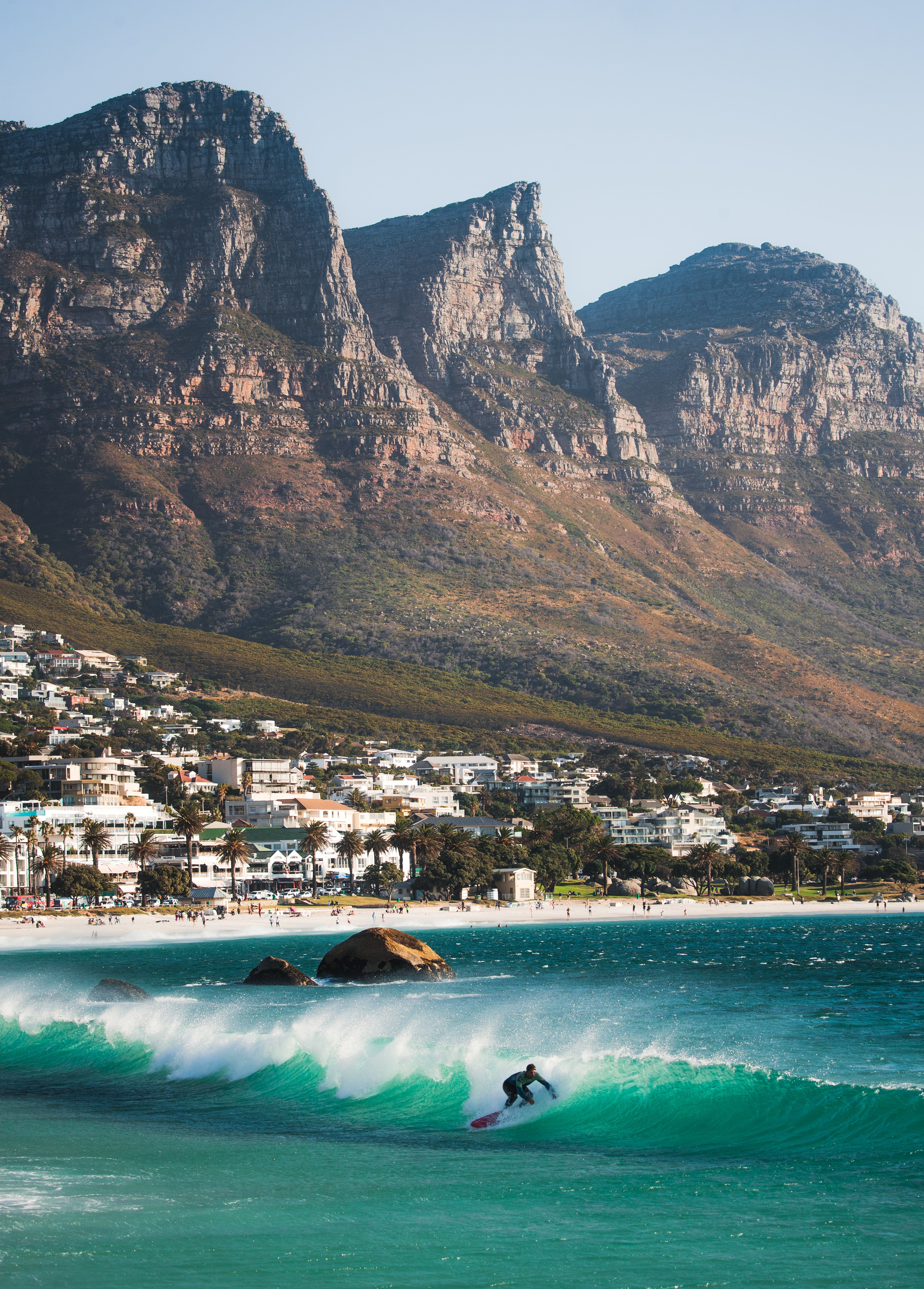 A surfer riding a wave in the turquoise waters of Camps Bay, Cape Town, with the stunning Twelve Apostles mountain range in the background