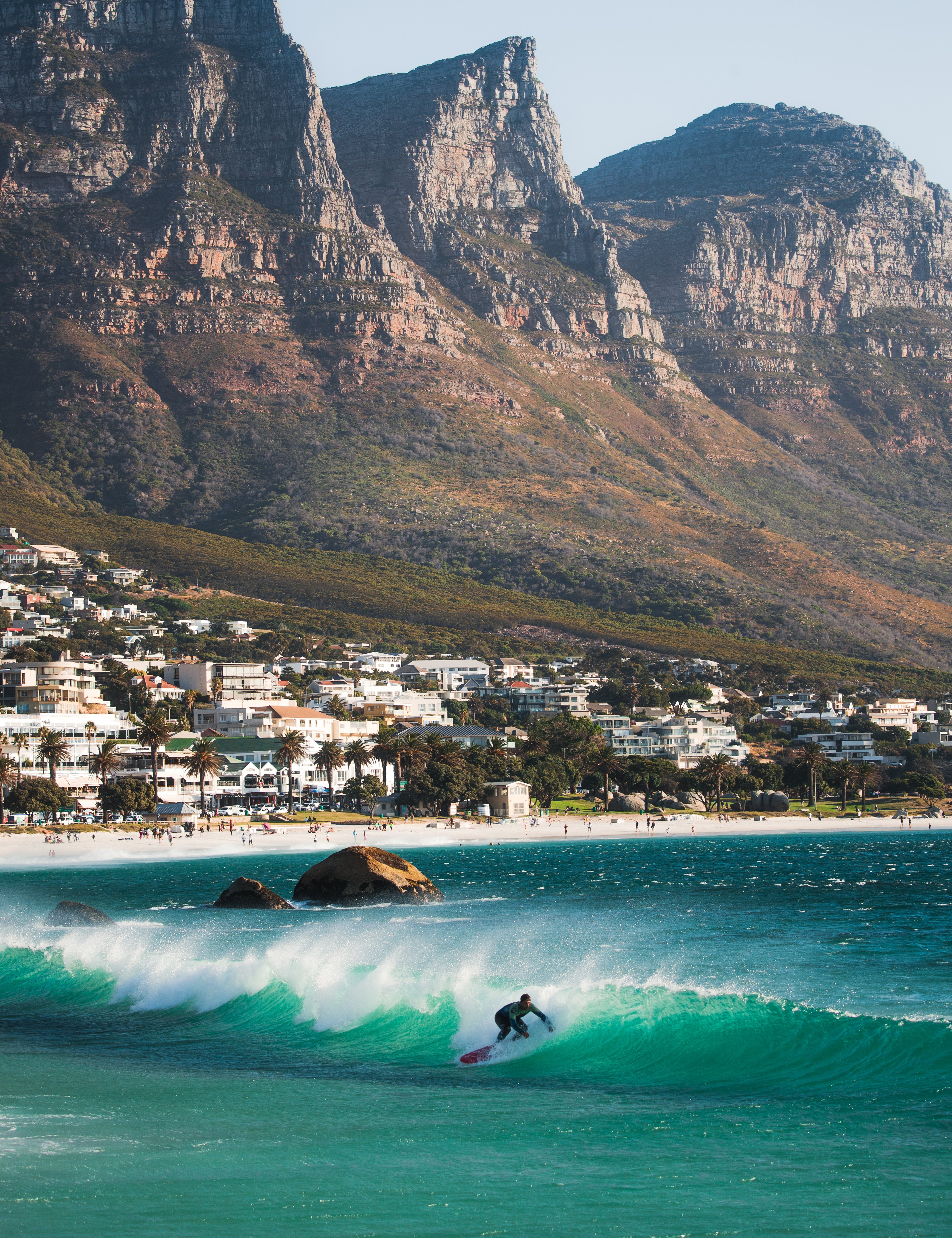 A surfer riding a wave in the turquoise waters of Camps Bay, Cape Town, with the stunning Twelve Apostles mountain range in the background