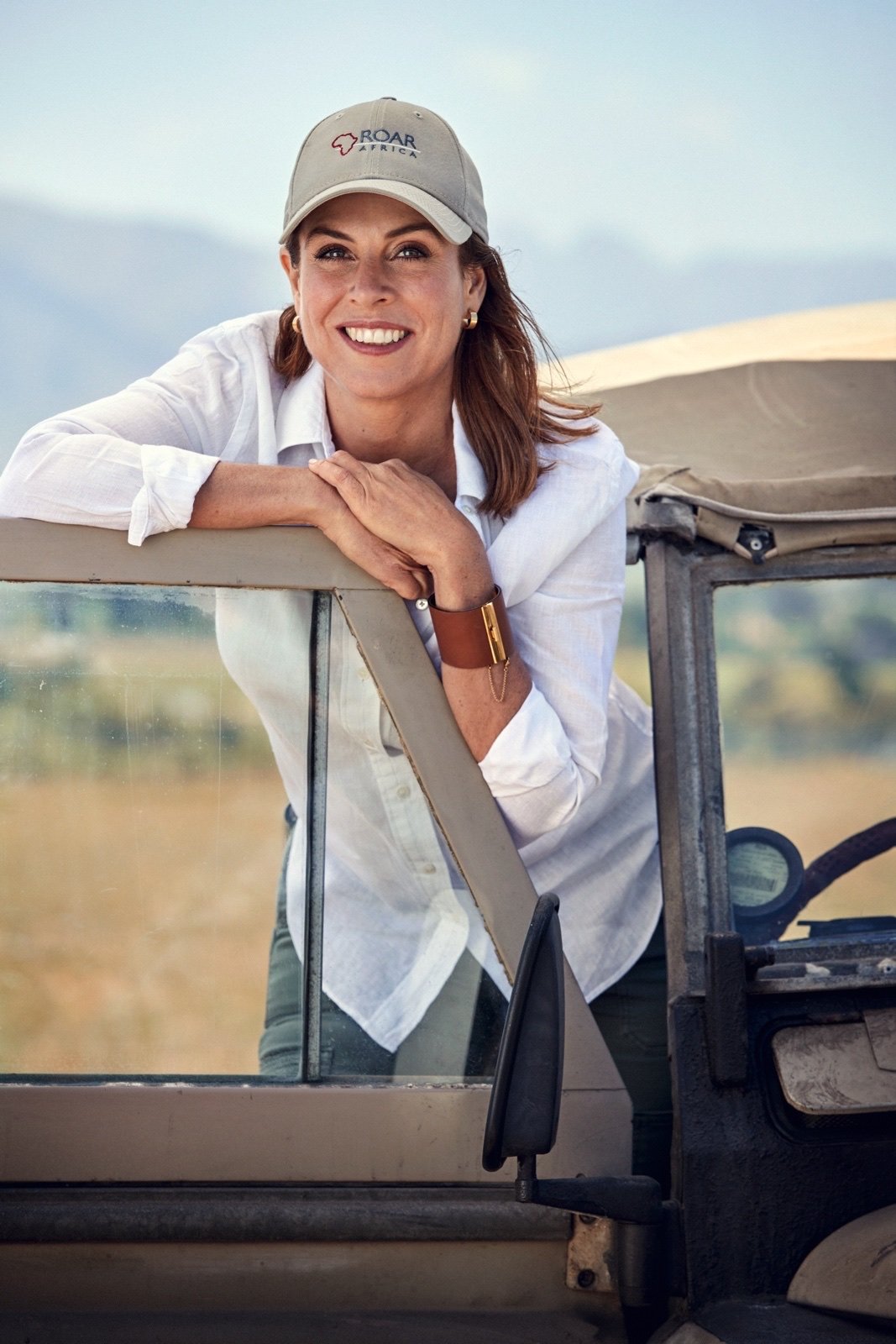 Portrait of Deborah Calmeyer, founder of ROAR AFRICA, in a ROAR AFRICA cap leaning on a safari vehicle in the African bush