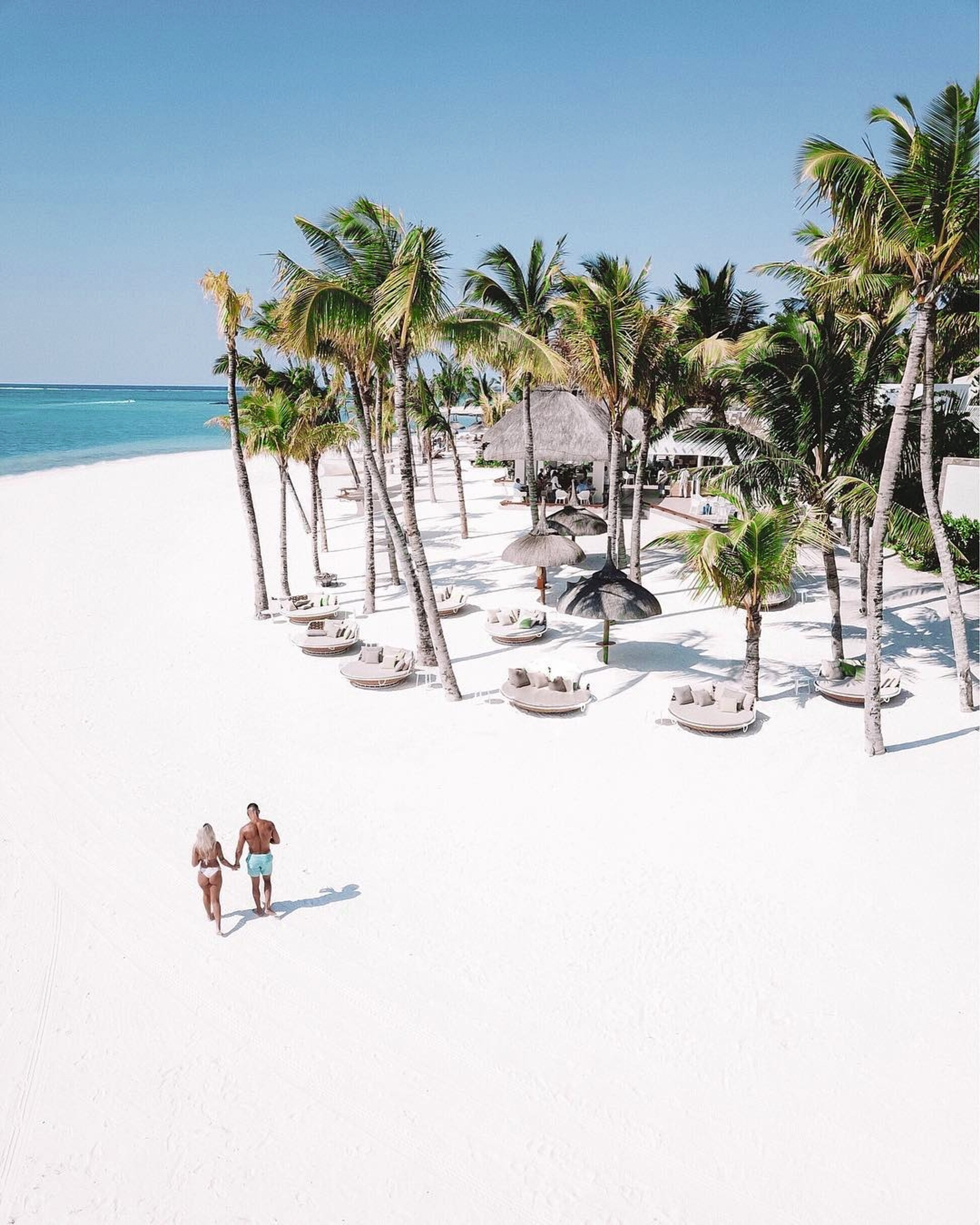 Couple walking on a white sandy beach lined with palm trees and lounge areas