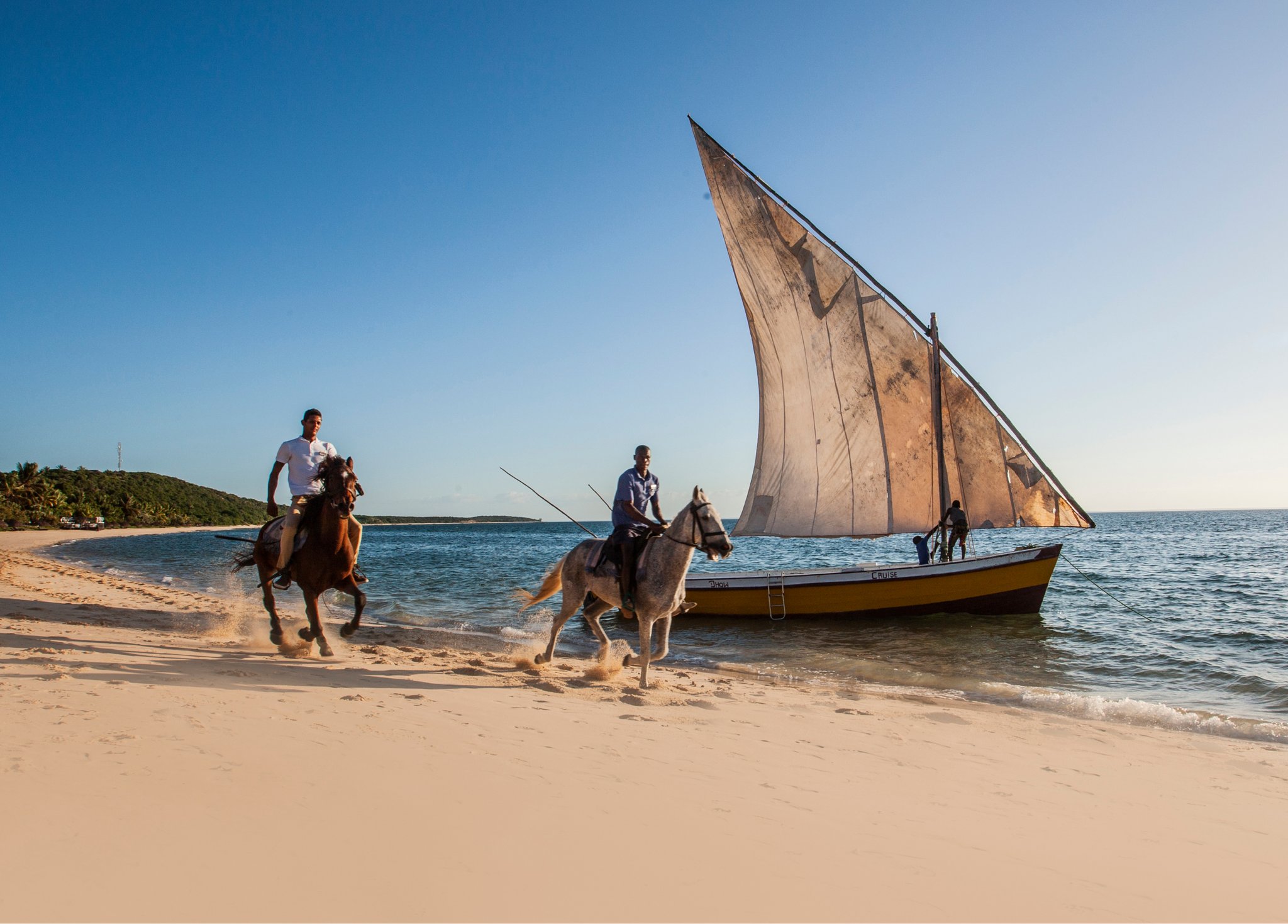 Two people riding horses on a beach near a sailboat