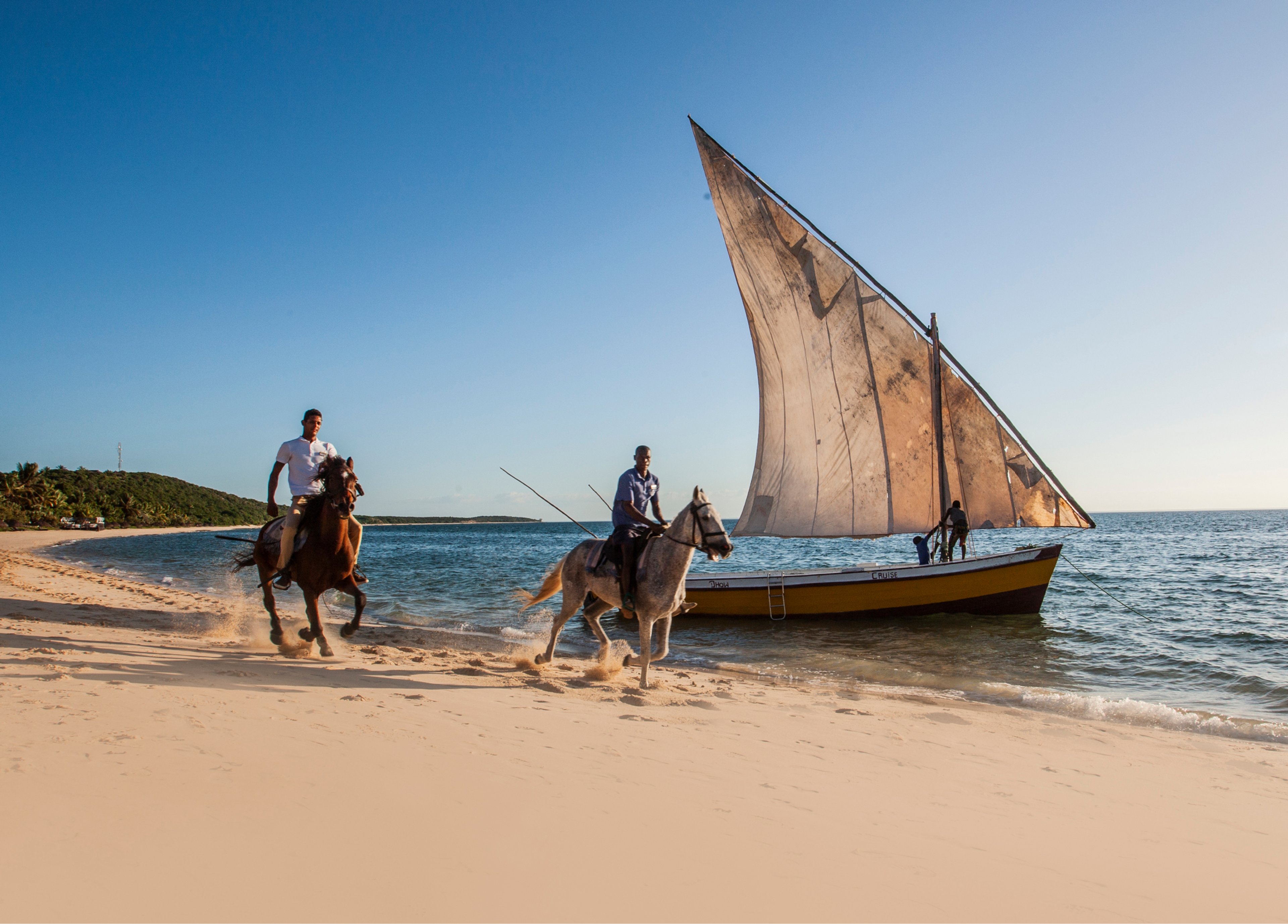 Two people riding horses on a beach near a sailboat