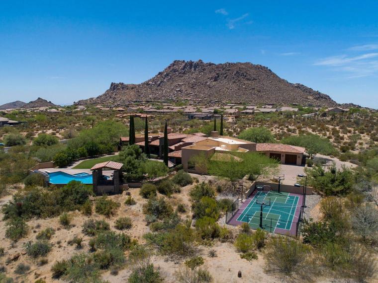 Aerial view of a desert home with a pool and private sport court, a large rounded rock mountain rising behind it