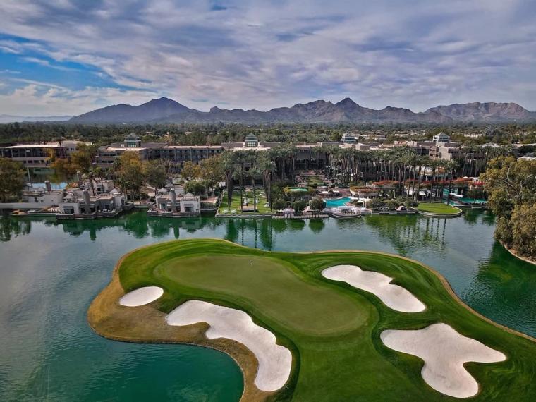 Aerial view of an island green ringed by bunkers and water, a resort and palm groves behind it and a mountain range beyond