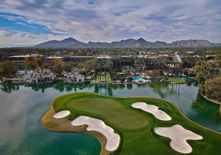 Aerial view of an island green ringed by bunkers and water, a resort and palm groves behind it and a mountain range beyond