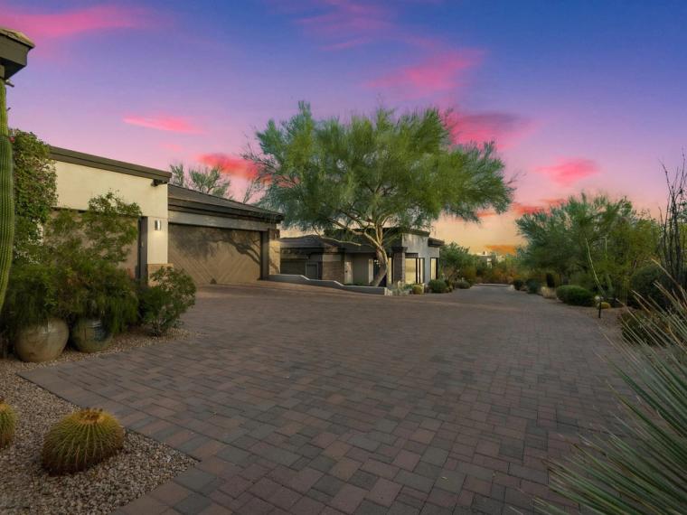 Contemporary desert home behind a wide paver motor court at dusk, barrel cactus and an ocotillo in the foreground under a pink and violet sky