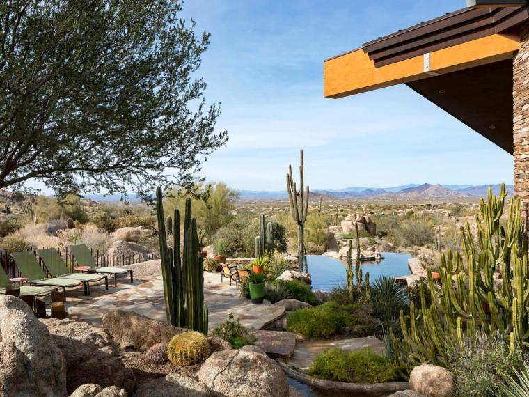 View from a shaded stone-and-timber patio out over cactus planting and a pond to open desert and distant hills